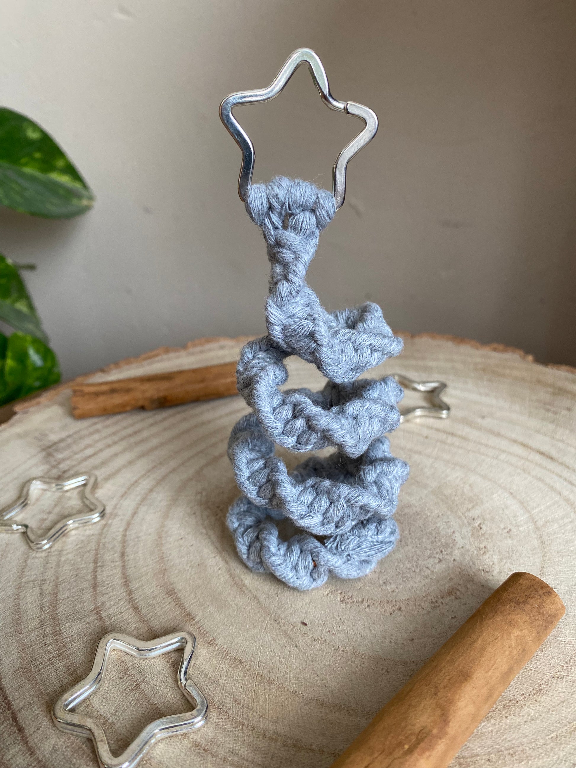 Close up image of a freestanding spiral Christmas tree ornament made with macrame knots in  grey yarn with a silver star on top, displayed on a wooden surface with metal stars and cinnamon sticks for decoration.