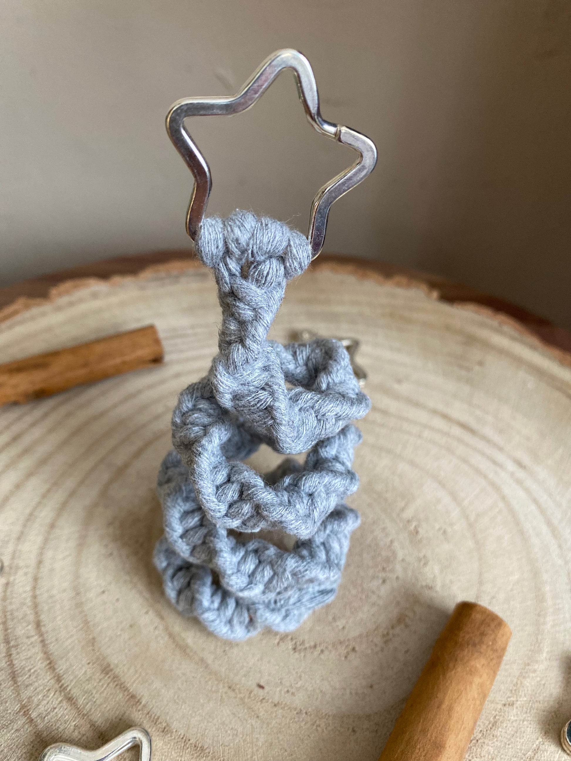 Close up of a freestanding spiral Christmas tree ornament made with macrame knots in grey yarn with a silver star on top, displayed on a wooden surface with cinnamon sticks for decoration.