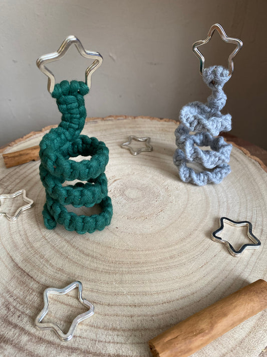 Two freestanding spiral Christmas tree ornaments made with macrame knots in green and grey colours with a silver star on top, displayed on a wooden surface with metal stars and cinnamon sticks for decoration.