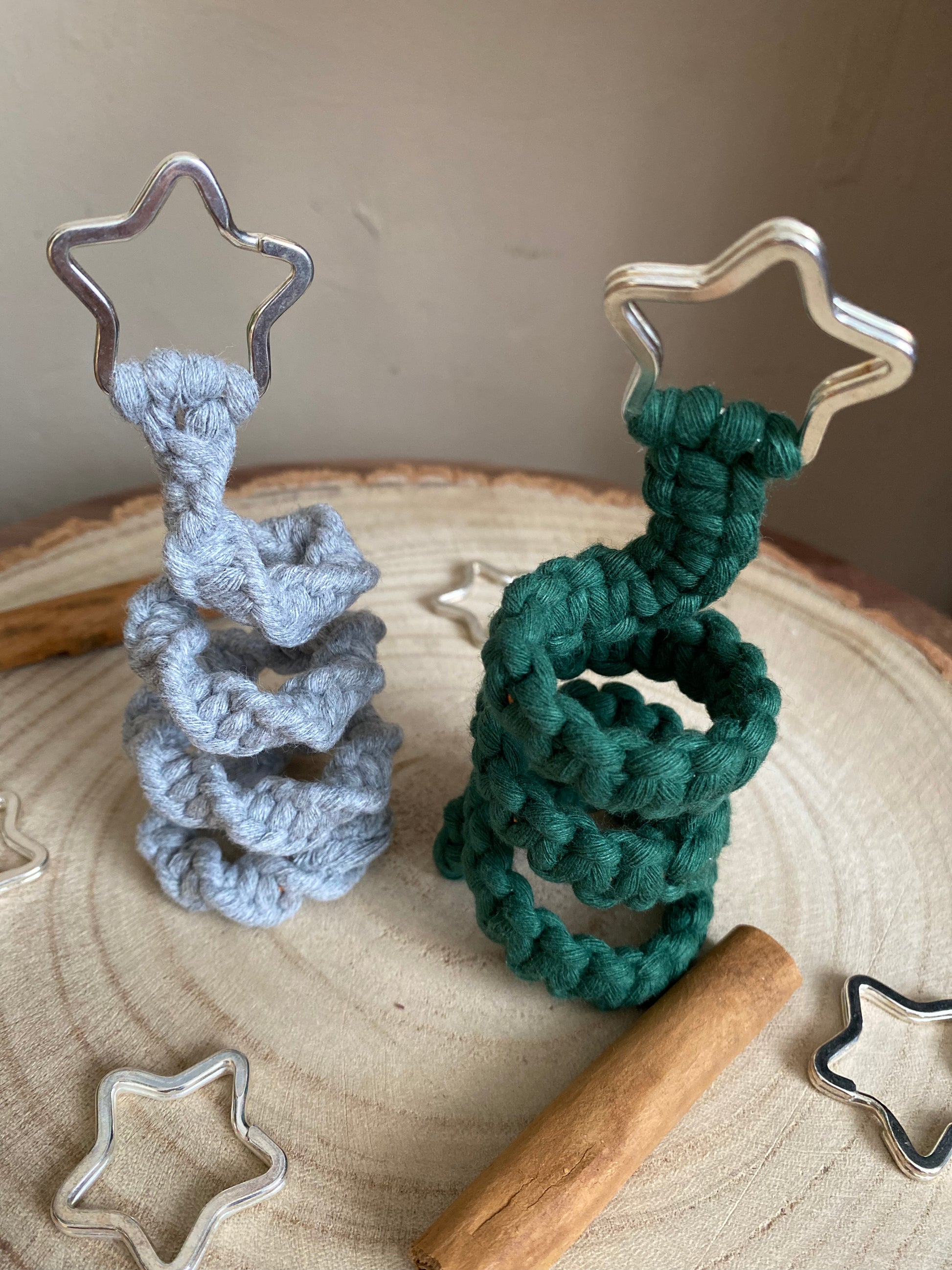 Close up image of two freestanding spiral Christmas tree ornaments made with macrame knots in grey and green yarn with a silver star on top of each, displayed on a wooden surface with metal stars and cinnamon sticks for decoration.