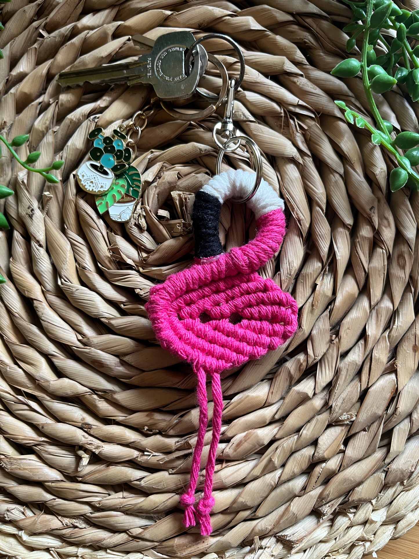 Handmade macrame keychain in the shape of a flamingo, made using fuchsia pink recycled cotton, on a metal split ring with lobster clasp. The keyring is displayed on a round wicker surface next to some green plant vines.