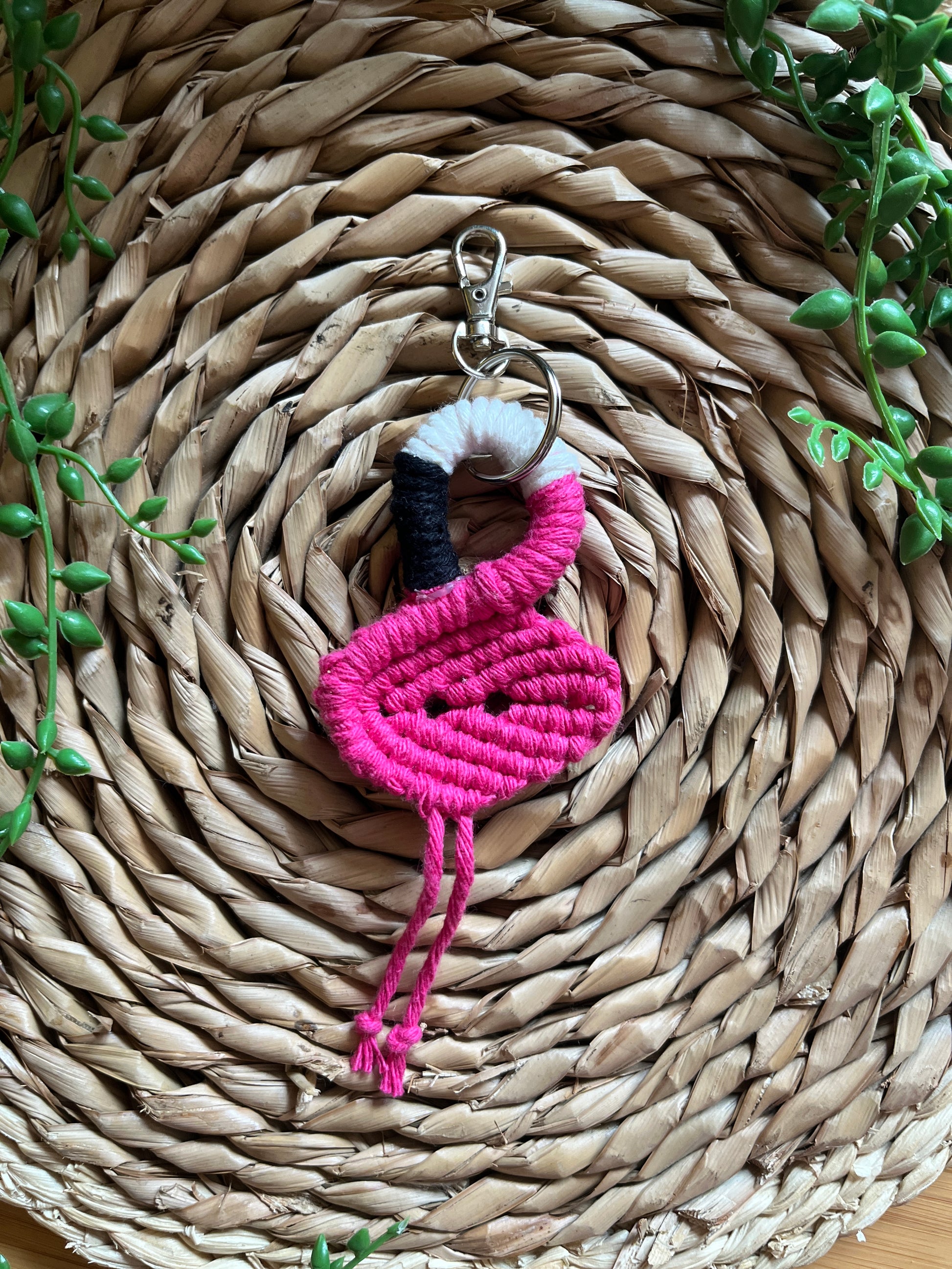 Handmade macrame keychain in the shape of a flamingo, made using fuchsia pink recycled cotton, on a metal split ring with lobster clasp. The keyring is displayed on a round wicker surface next to some green plant vines.