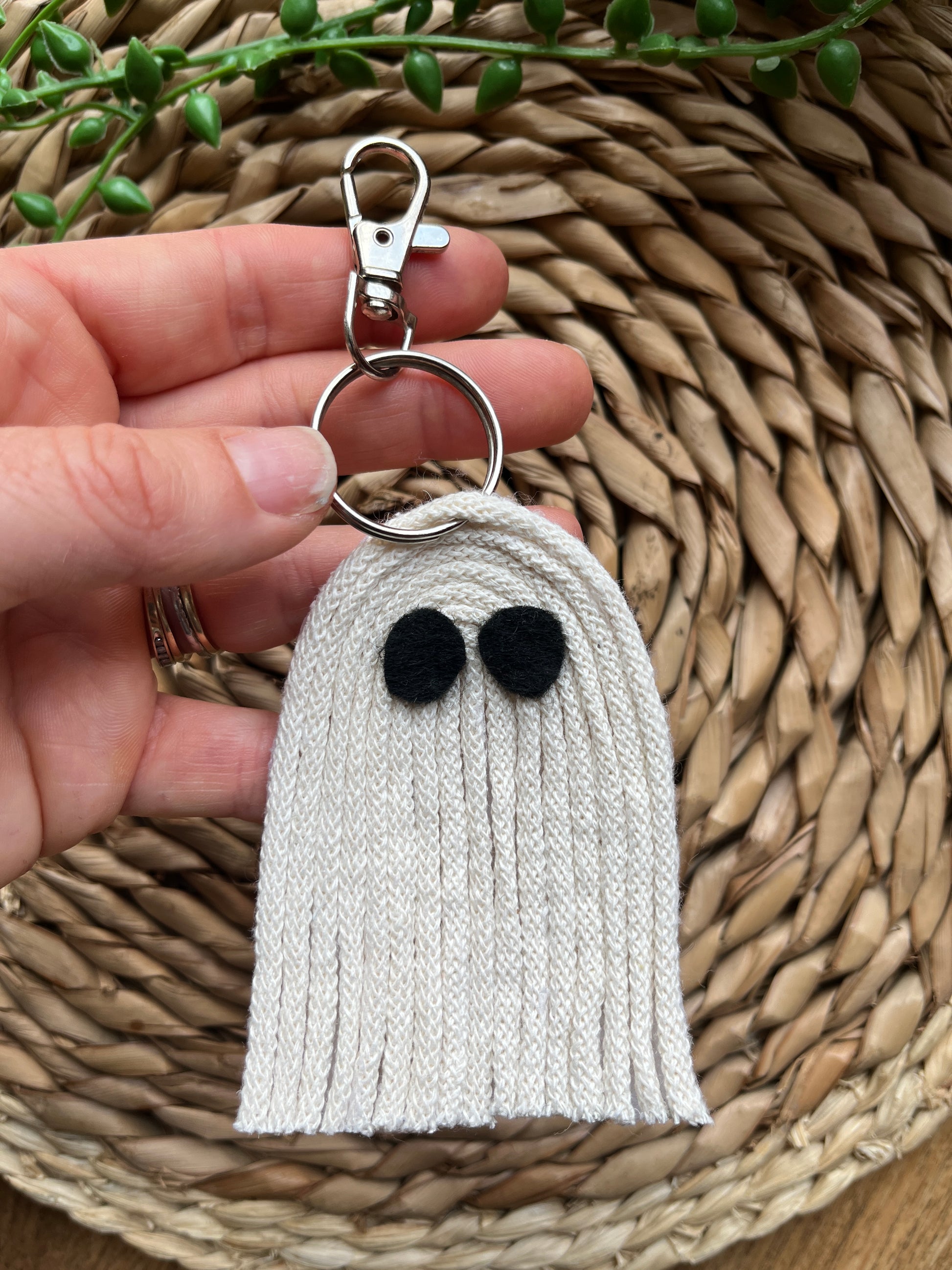 White ghost-shaped keychain with black felt eyes, held by a hand on a woven surface.