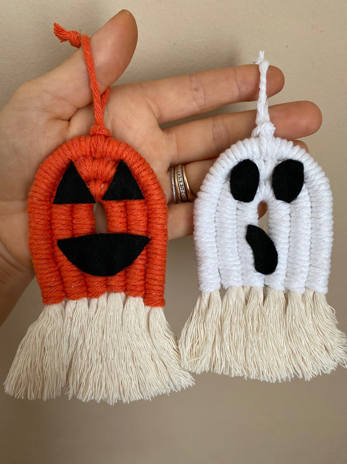 A hand holding macrame ghost and pumpkin ornaments, made with orange and white yarns with black felt face details and long natural tassels at the bottom.