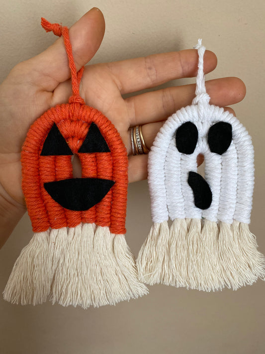 A hand holding macrame ghost and pumpkin ornaments, made with orange and white yarns with black felt face details and long natural tassels at the bottom.