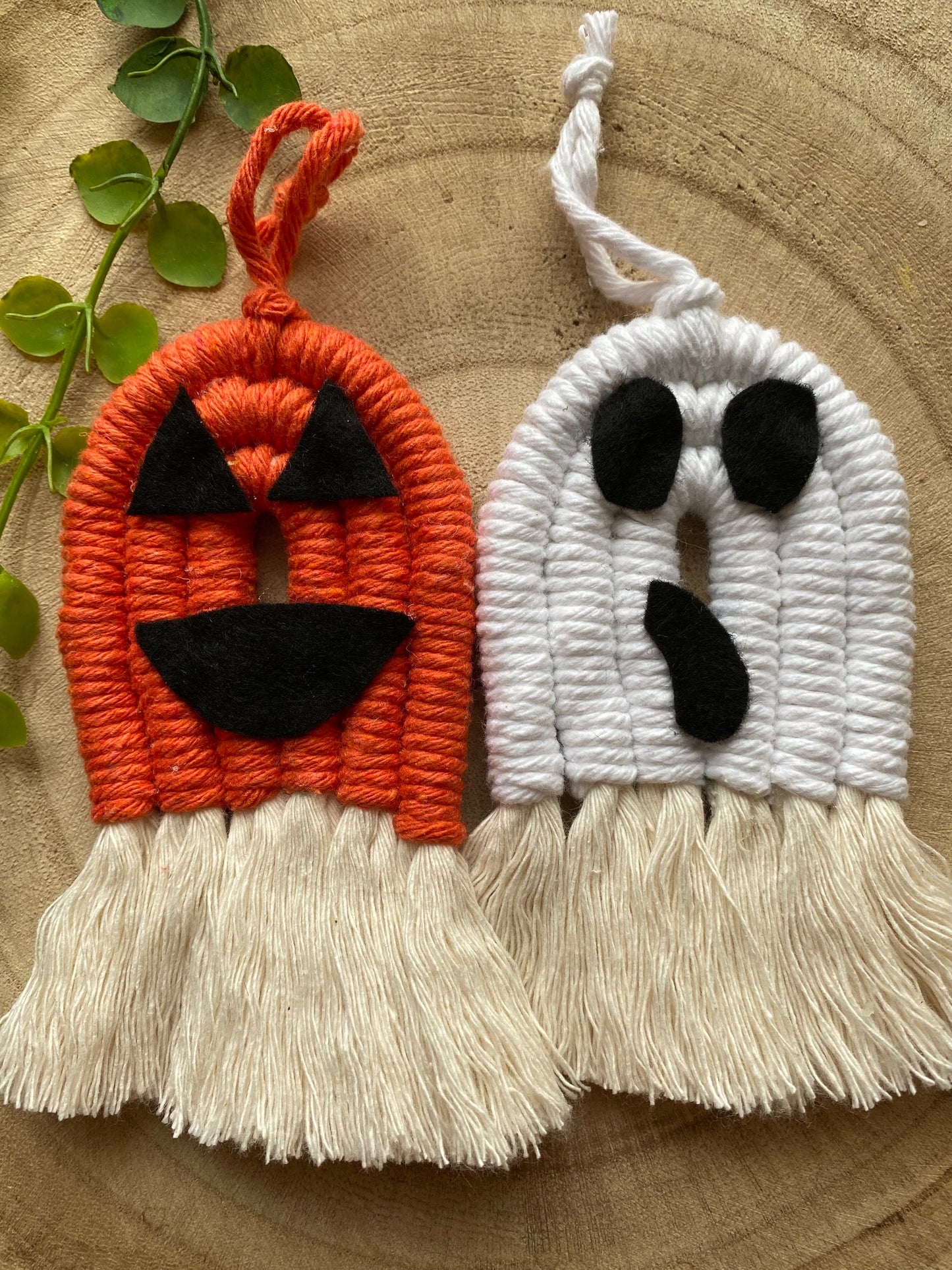 A pair of macrame ghost and pumpkin ornaments, made with orange and white yarns with black felt face details and long natural tassels at the bottom, displayed next to each other on a wooden board.
