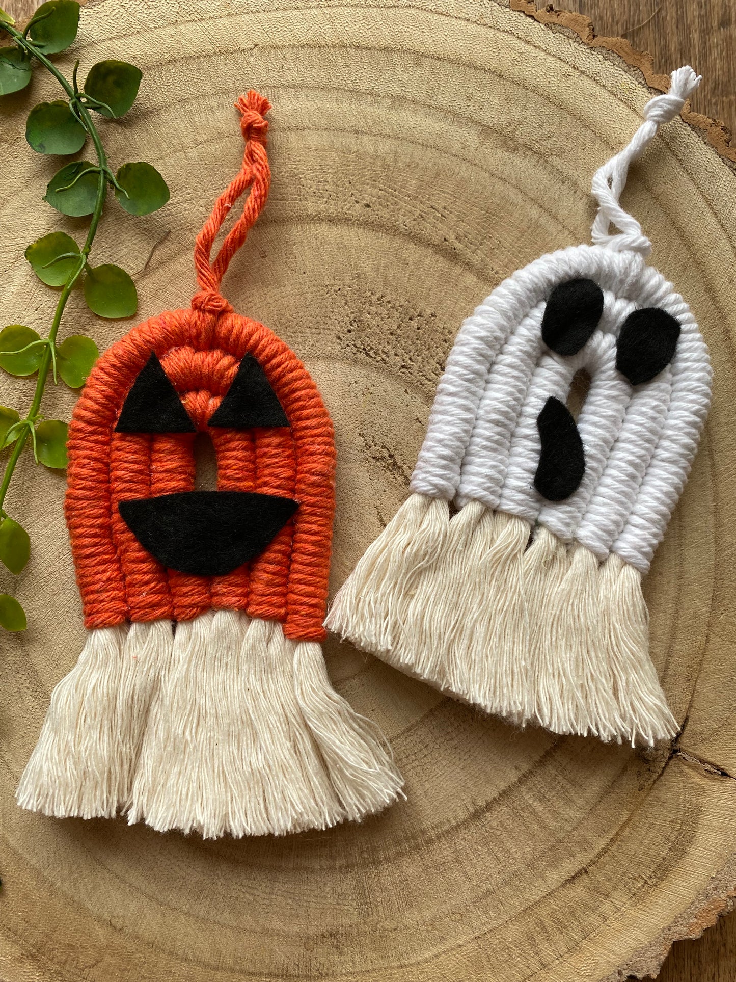A set of macrame ghost and pumpkin ornaments, made with orange and white yarns with black felt face details and long natural tassels at the bottom on a wooden board.