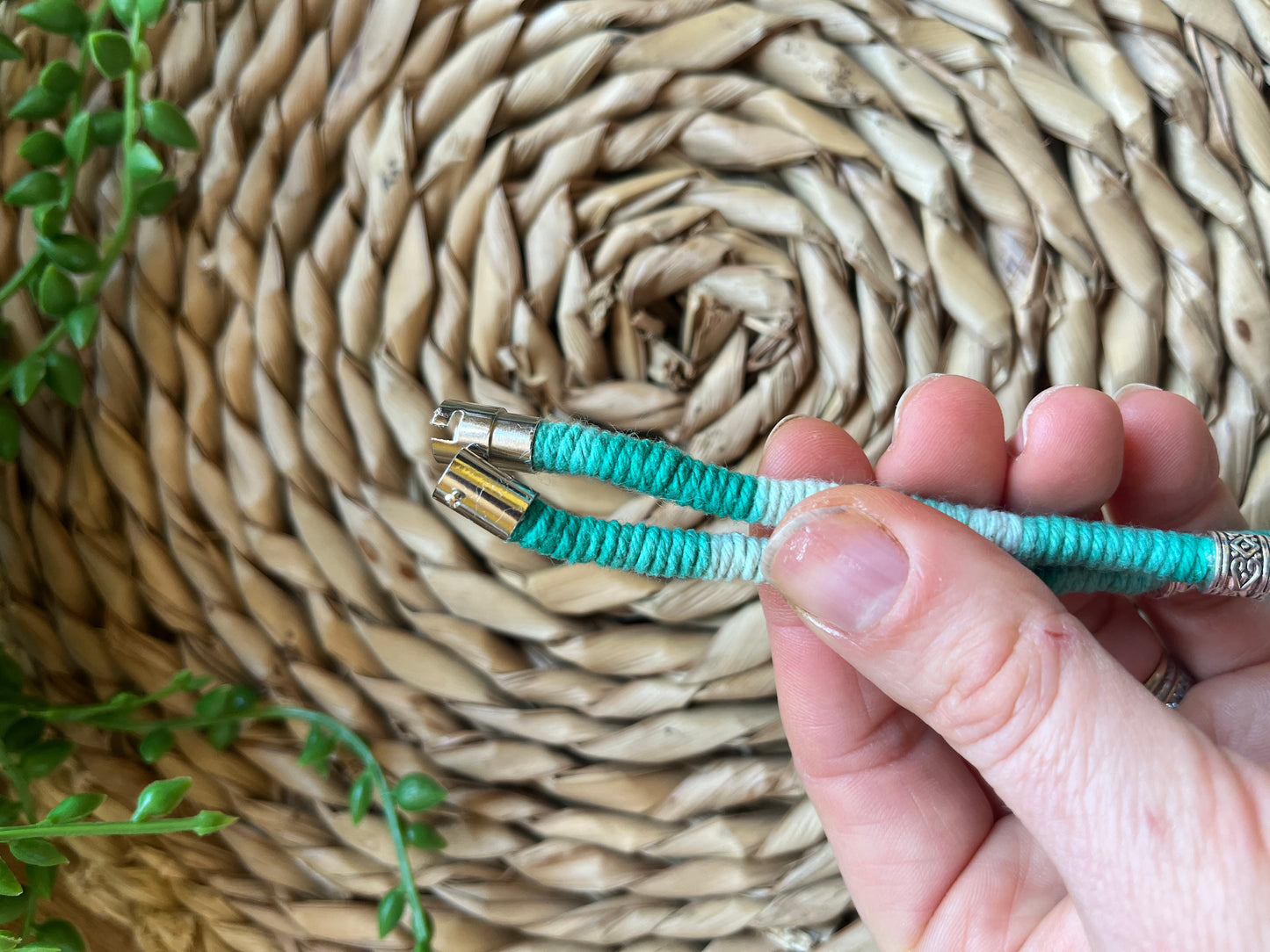 A hand holding a handmade macrame surfer style short boho necklace, handmade using eco friendly green variegated yarn, and 3 Celtic style spacer beads. Displayed on a wicker mat next to some green plant vines. The image focuses on the magnetic clasp fastening on the necklace.