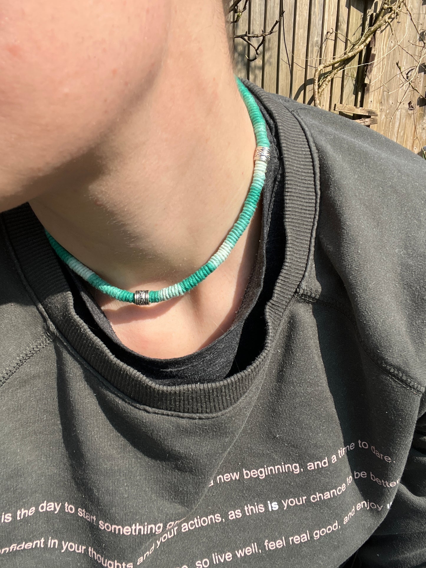 Handmade macrame surfer style short boho necklace, handmade using eco friendly green variegated yarn, and 3 Celtic style spacer beads. The necklace is being worn by a female, wearing a dark green jumper, outside, with a wooden fence visible in the background.