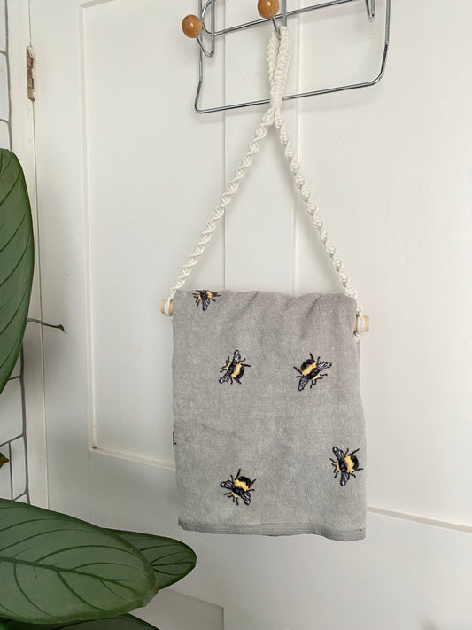 A grey hand towel with bee design hangs on a macrame hand towel holder, made with natural coloured recycled cotton, which is hanging on the back of a white painted door.