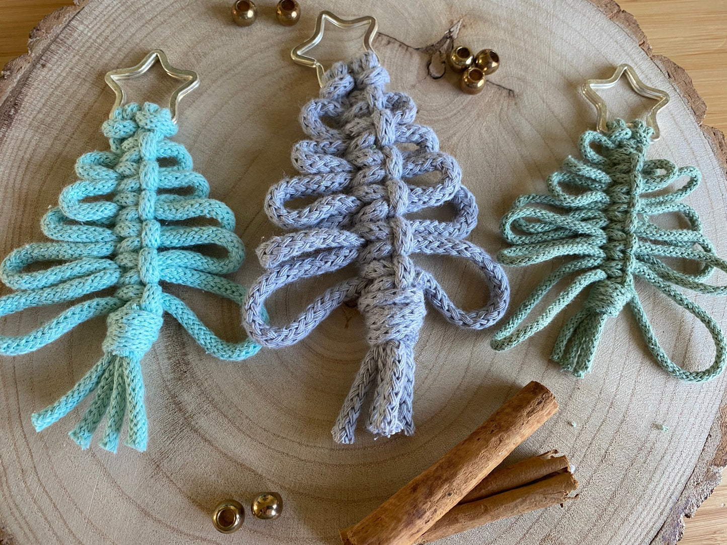 Three knotted yarn hanging Christmas tree decorations by Macra-Made-With-Love in pastel shades (mint green, light blue, and light grey) lie on a wooden surface with a tree ring pattern. Each ornament features a star at the top. Nearby, cinnamon sticks and small bronze jingle bells help create an eco-friendly atmosphere for your handmade holiday decor.