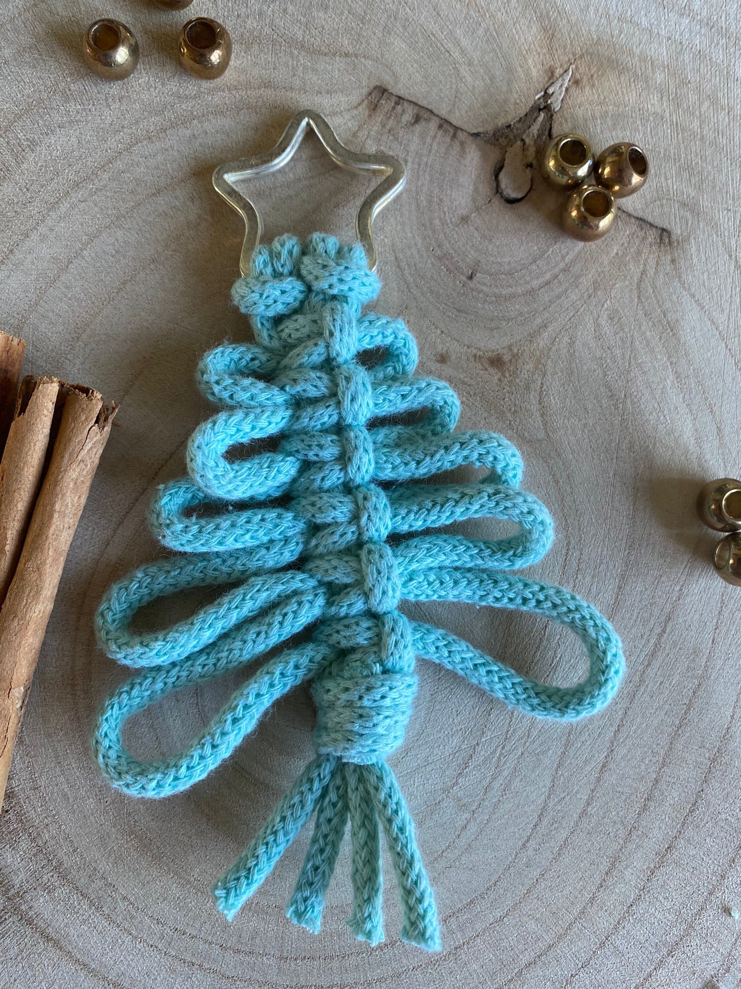 A blue Macra-Made-With-Love Knotted Yarn Hanging Christmas Tree Decoration with a silver star at the top is intricately woven using recycled cotton yarns and lies on a wooden surface. Cinnamon sticks and small golden bells are scattered around, creating an eco-friendly holiday decor setting.