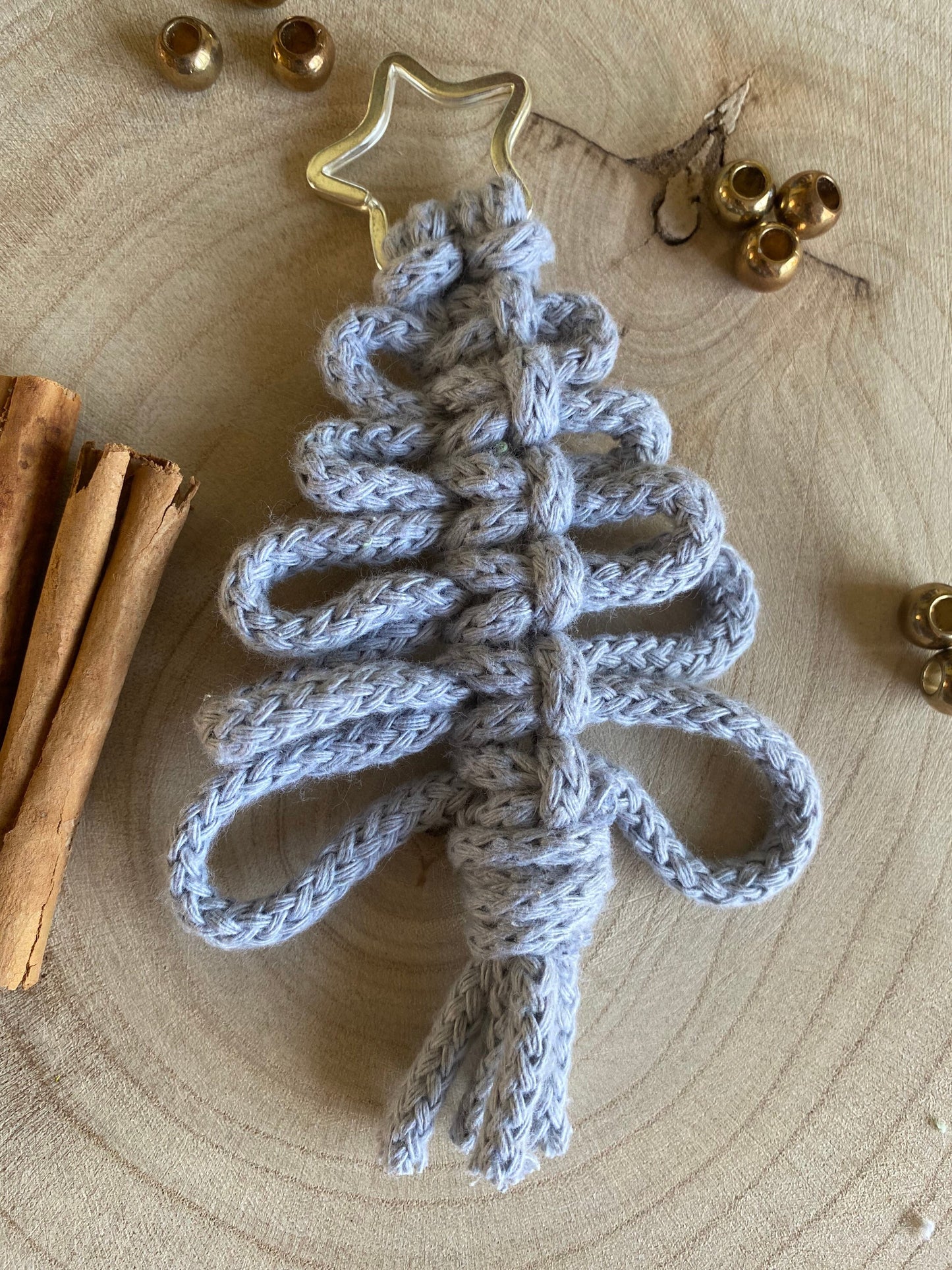 A handmade holiday decor piece, the Knotted Yarn Hanging Christmas Tree Decoration from Macra-Made-With-Love is displayed on a wooden surface. Crafted from light gray yarn and topped with a golden star, it features eco-friendly decorations like cinnamon sticks and small gold bells arranged around the tree.