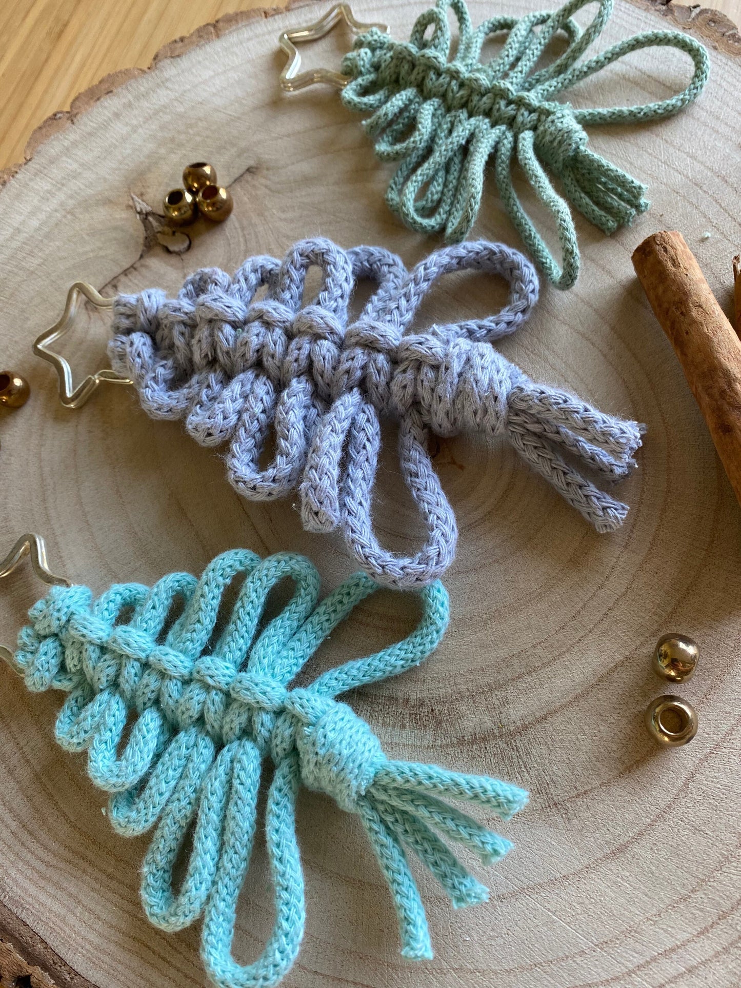 A circular wooden surface showcases three Knotted Yarn Hanging Christmas Tree Decorations by Macra-Made-With-Love in light green, pastel blue, and gray, each adorned with a star-shaped top. These handmade holiday decor items are enhanced by small golden bells and a cinnamon stick, creating an eco-friendly and cozy festive atmosphere.