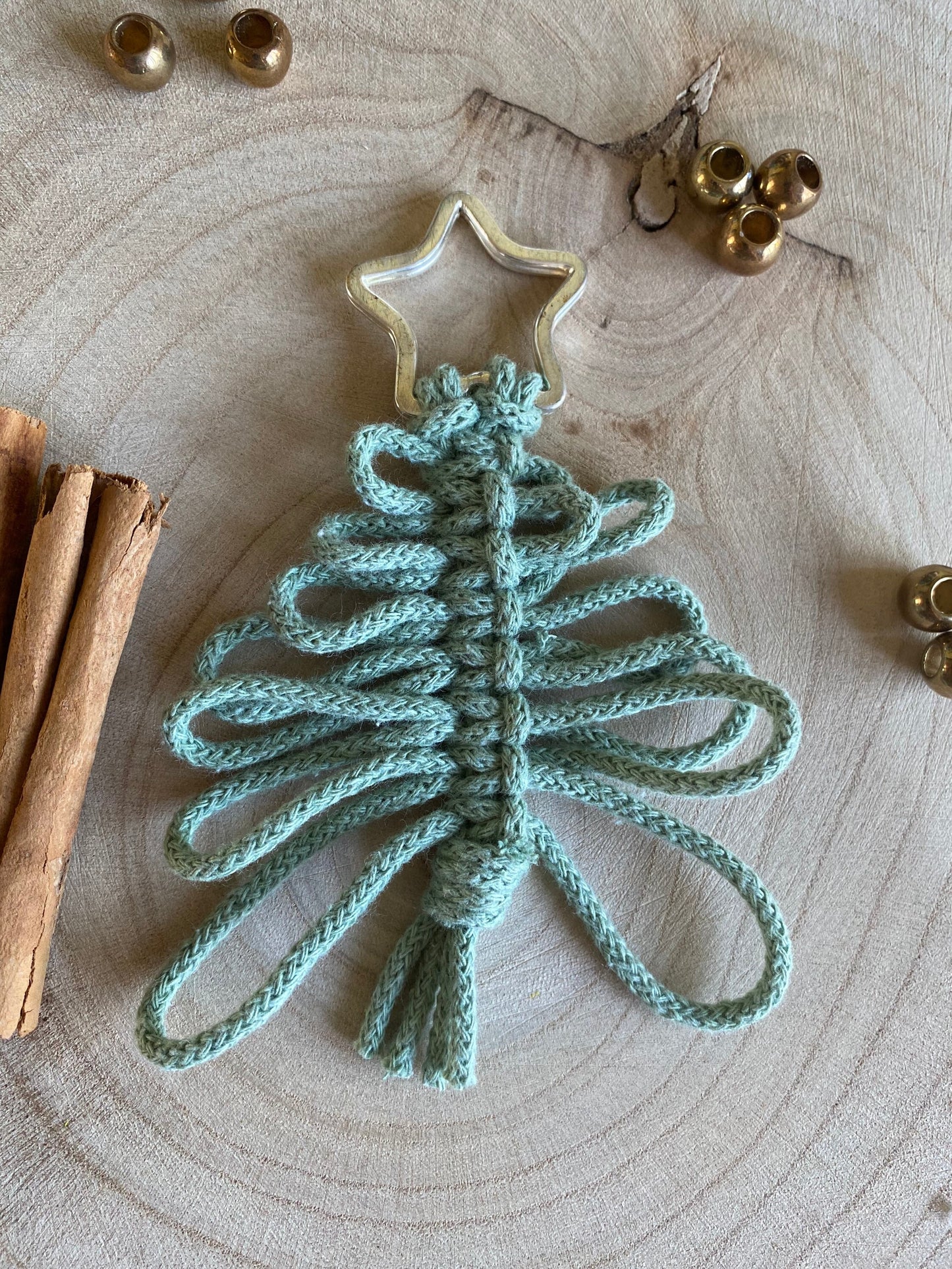 Introducing the Knotted Yarn Hanging Christmas Tree Decoration from Macra-Made-With-Love, this handmade holiday ornament is crafted from light blue yarn and shaped like a tree with looped branches and a knotted base. It features a golden star on top and is displayed on a circular wooden surface decorated with cinnamon sticks and small bells.