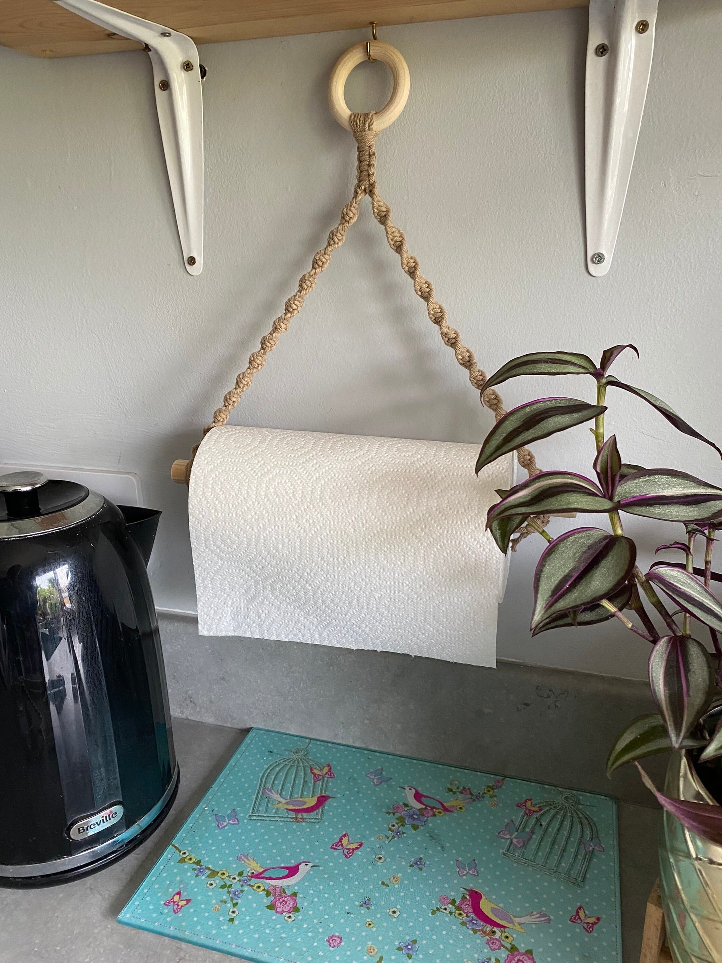 A roll of paper towels hangs from a Macra-Made-With-Love Kitchen roll holder attached to a wall under a wooden shelf, beside a black electric kettle. Below the paper towels, there's a colorful mat with birds and flowers, and to the right, a potted plant with variegated leaves.
