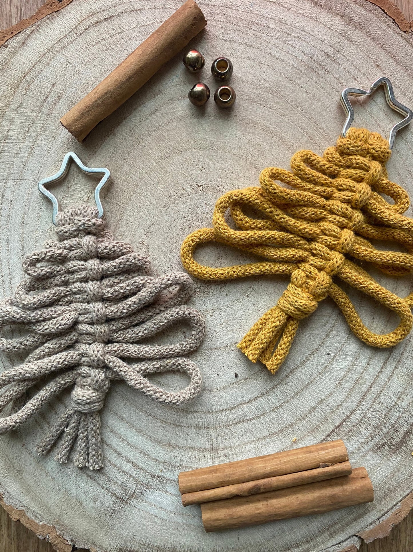 Two eco-friendly Knotted Yarn Hanging Christmas Tree Decorations from Macra-Made-With-Love on a wooden surface. One is beige and the other is yellow, both with loops forming the tree shape and a star-shaped top. Three cinnamon sticks and four golden beads are scattered around these handmade holiday decor pieces.