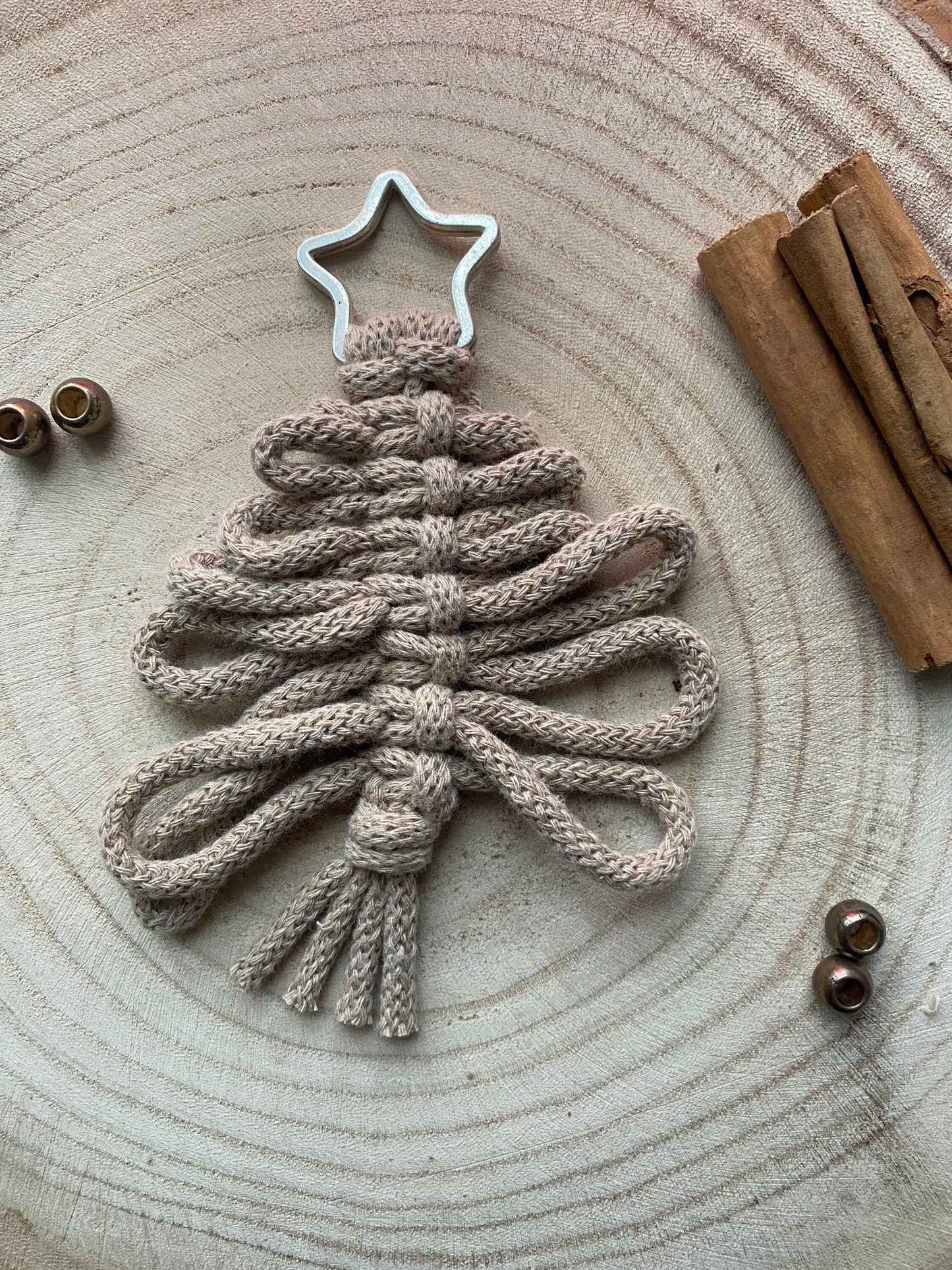 Introducing Macra-Made-With-Love's Knotted Yarn Hanging Christmas Tree Decorations, featuring eco-friendly materials in a charming zigzag pattern and crowned with a silver star. Set against a wooden background accented with cinnamon sticks and small metal beads, the natural textures and colors offer a cozy, rustic ambiance perfect for handmade holiday decor.