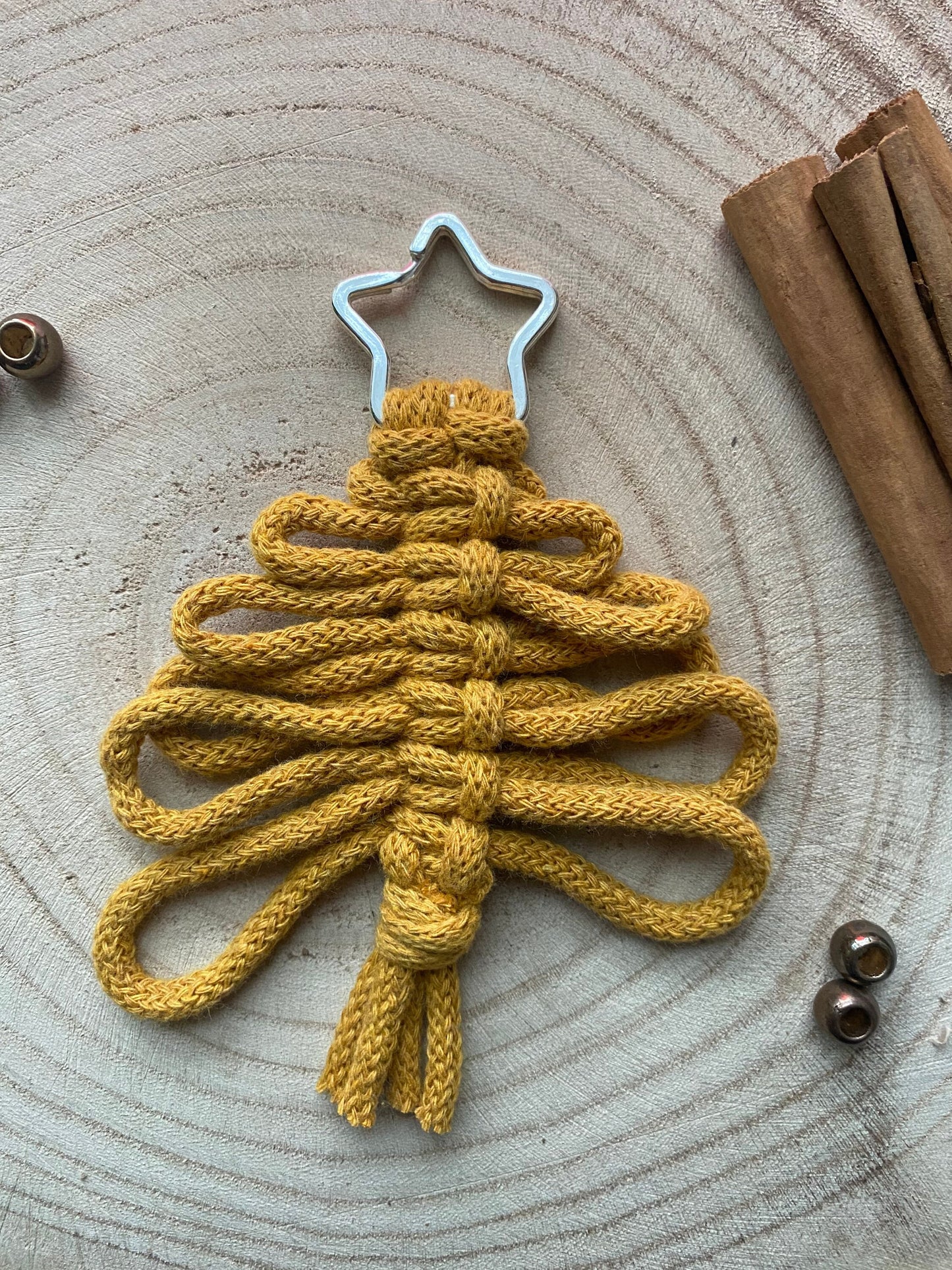 A handmade holiday decor piece, the Knotted Yarn Hanging Christmas Tree Decorations by Macra-Made-With-Love are crafted from yellow macrame yarn, intricately looped in layers to form a charming tree shape. Topped with a metallic star and set on a wooden surface with cinnamon sticks and decorative beads nearby, they add an eco-friendly charm to your festive decorations.