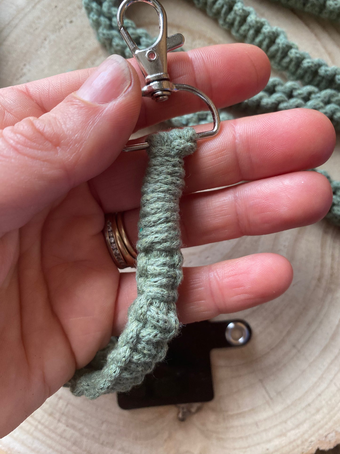 A close-up of a hand holding a green crocheted loop with a metal clip attached, part of the Recycled cotton crossbody phone strap by Macra-Made-With-Love. The person's thumb and two fingers are visible, adorned with rings. The background is a wooden surface.