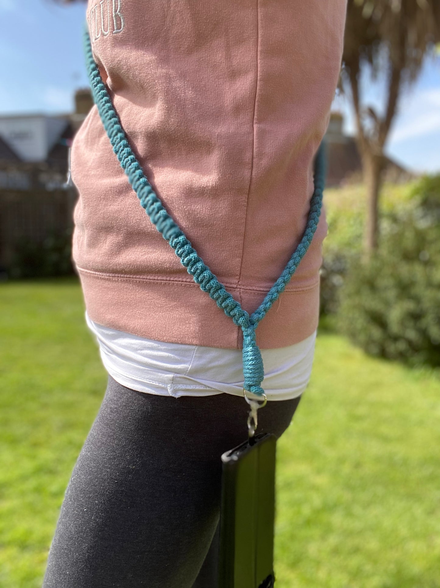 A person in a pink hoodie and gray leggings is seen wearing an eco-friendly, handmade Macra-Made-With-Love Recycled cotton crossbody phone strap around their waist, with a black pouch attached. They are outdoors in a grassy area with some houses and trees in the background.