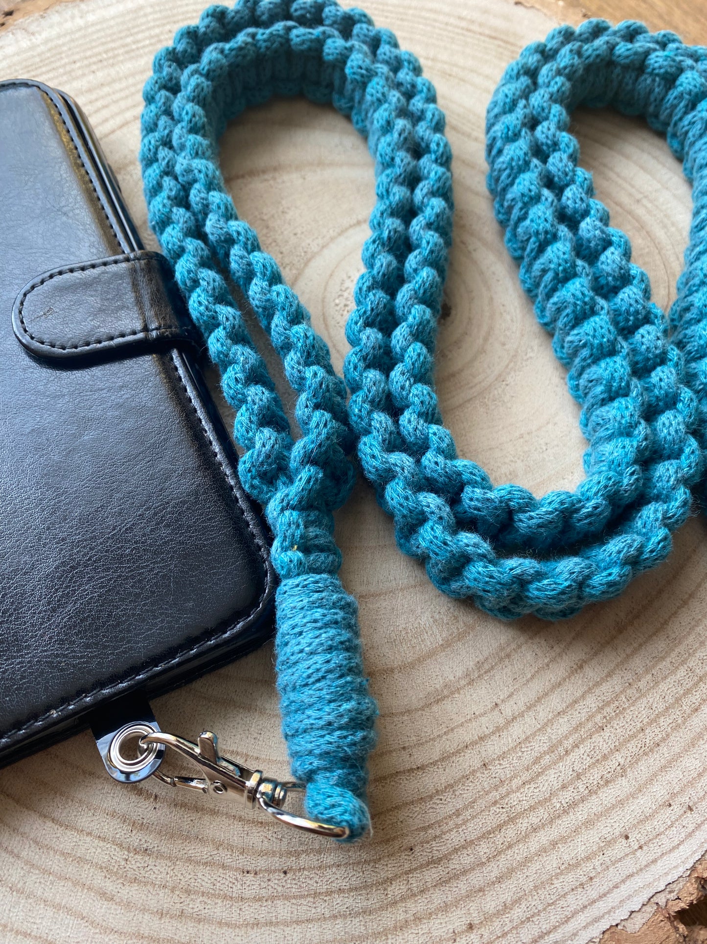 A teal, braided rope lanyard with a silver clip lies on a wooden surface next to a black leather wallet with a snap closure. This Recycled cotton crossbody phone strap by Macra-Made-With-Love is coiled in a loose, looping pattern.
