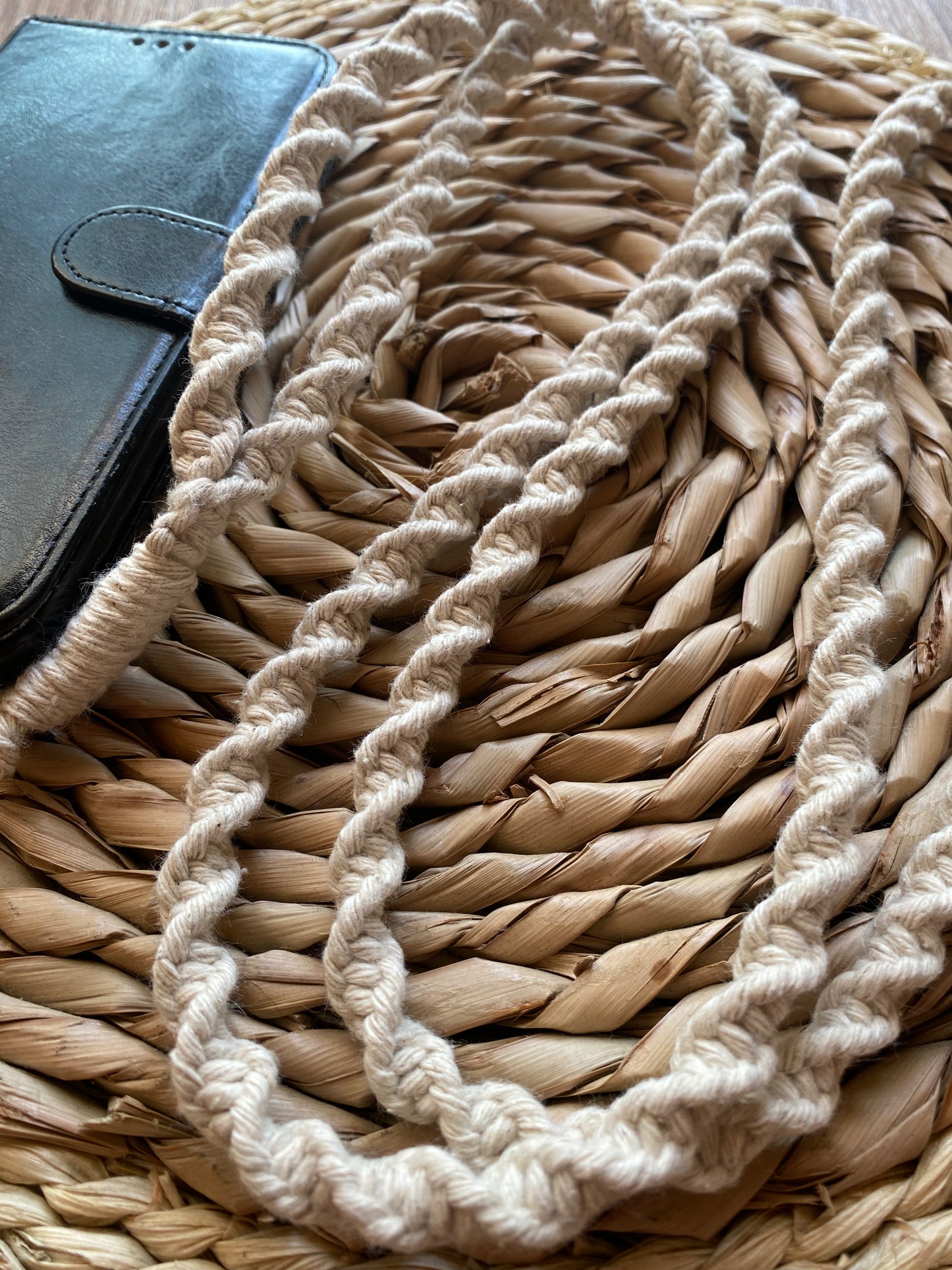 A black wallet lies on a woven straw mat. Draped across the mat is a long, intricately twisted beige rope, handmade into an eco-friendly Recycled cotton crossbody phone strap by Macra-Made-With-Love. The braided texture of the strap contrasts with the smooth leather surface of the wallet and the natural texture of the mat.