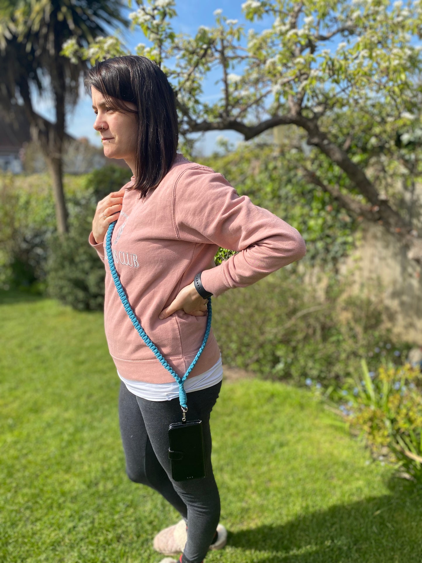 A woman stands in a garden, looking to her left. She wears a pink sweatshirt, dark gray leggings, and slippers. A Recycled cotton crossbody phone strap from Macra-Made-With-Love, eco-friendly and stylish, is slung over her shoulder with a black phone attached. A tree and shrubbery are visible in the background.