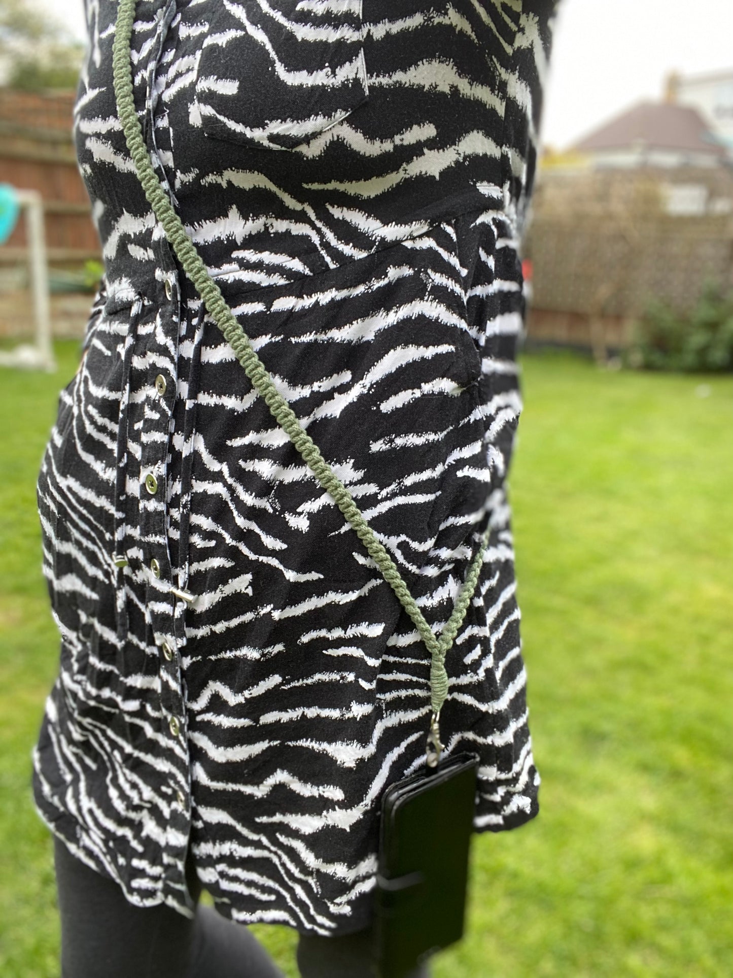 A person wearing a black and white zebra-patterned dress stands outdoors, with an eco-friendly Recycled cotton crossbody phone strap by Macra-Made-With-Love hanging from their shoulder. The background features a green lawn, wooden fence, and a house visible in the distance.
