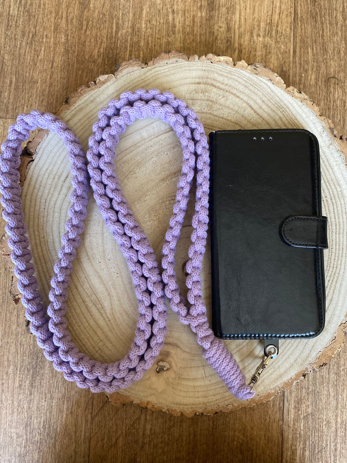 A black phone case with a front flap is attached to a long, handmade Recycled cotton crossbody phone strap by Macra-Made-With-Love. The strap is coiled neatly around the phone case and placed on a circular wooden surface with visible tree rings, set on a wooden background. This eco-friendly phone accessory combines style and functionality seamlessly.