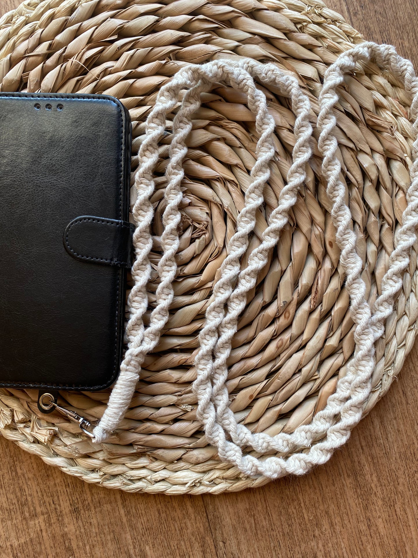 A black smartphone wallet case with a snap closure is paired with the Recycled cotton crossbody phone strap from Macra-Made-With-Love. The items are placed on a round woven placemat, which is set on a wooden surface.