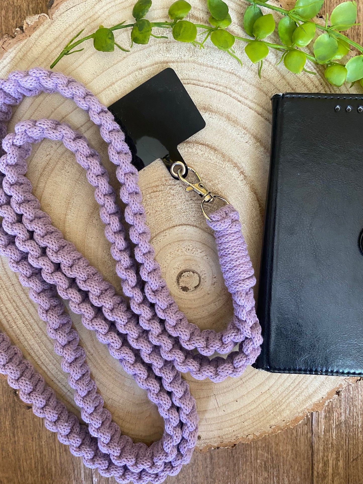 A chunky, crocheted lavender lanyard with a black ID holder is displayed on a wooden slice, surrounded by green leaves. Beside it lies an eco-friendly, handmade Recycled cotton crossbody phone strap by Macra-Made-With-Love and a closed black notebook with a dotted cover.