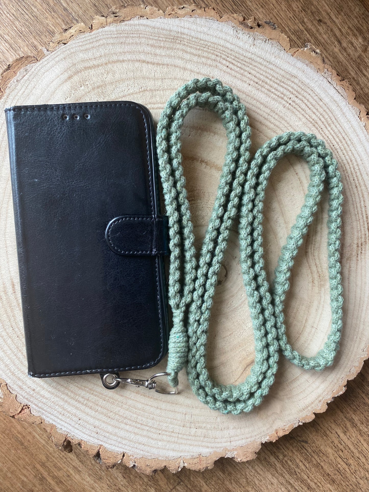 A black leather phone wallet and a green Recycled cotton crossbody phone strap by Macra-Made-With-Love are placed on a round wooden surface with bark edges, resembling a tree cross-section. The eco-friendly lanyard has a metal clasp at one end.