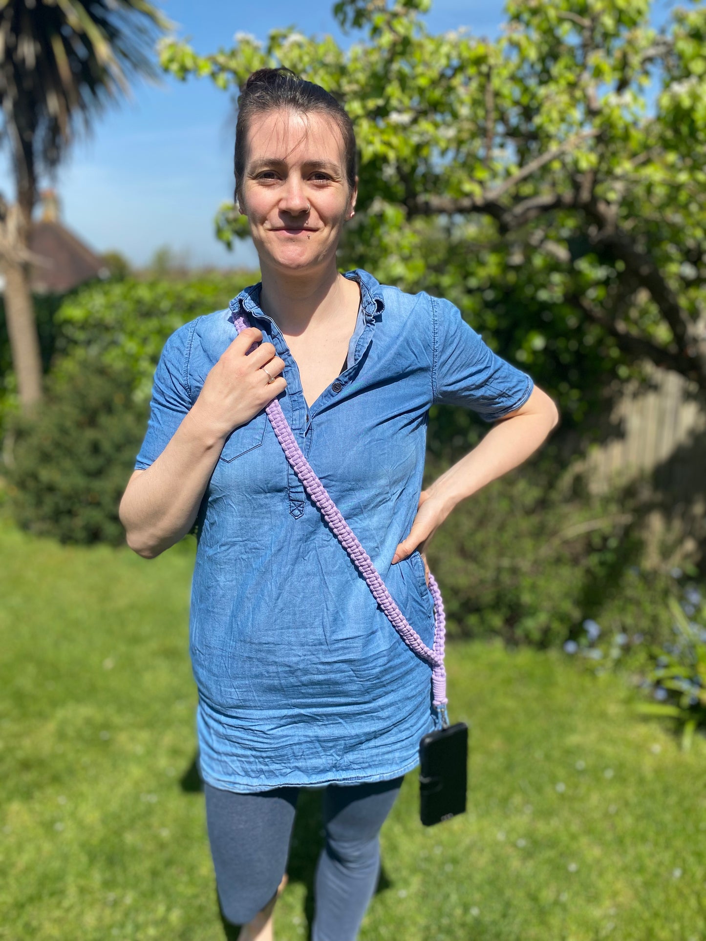 A person stands outdoors on a sunny day, wearing a blue tunic and gray leggings. They have a phone with a Macra-Made-With-Love recycled cotton crossbody phone strap over their shoulder. Green foliage, including a tree and bushes, is visible in the background. The person smiles slightly, with one hand on their hip.