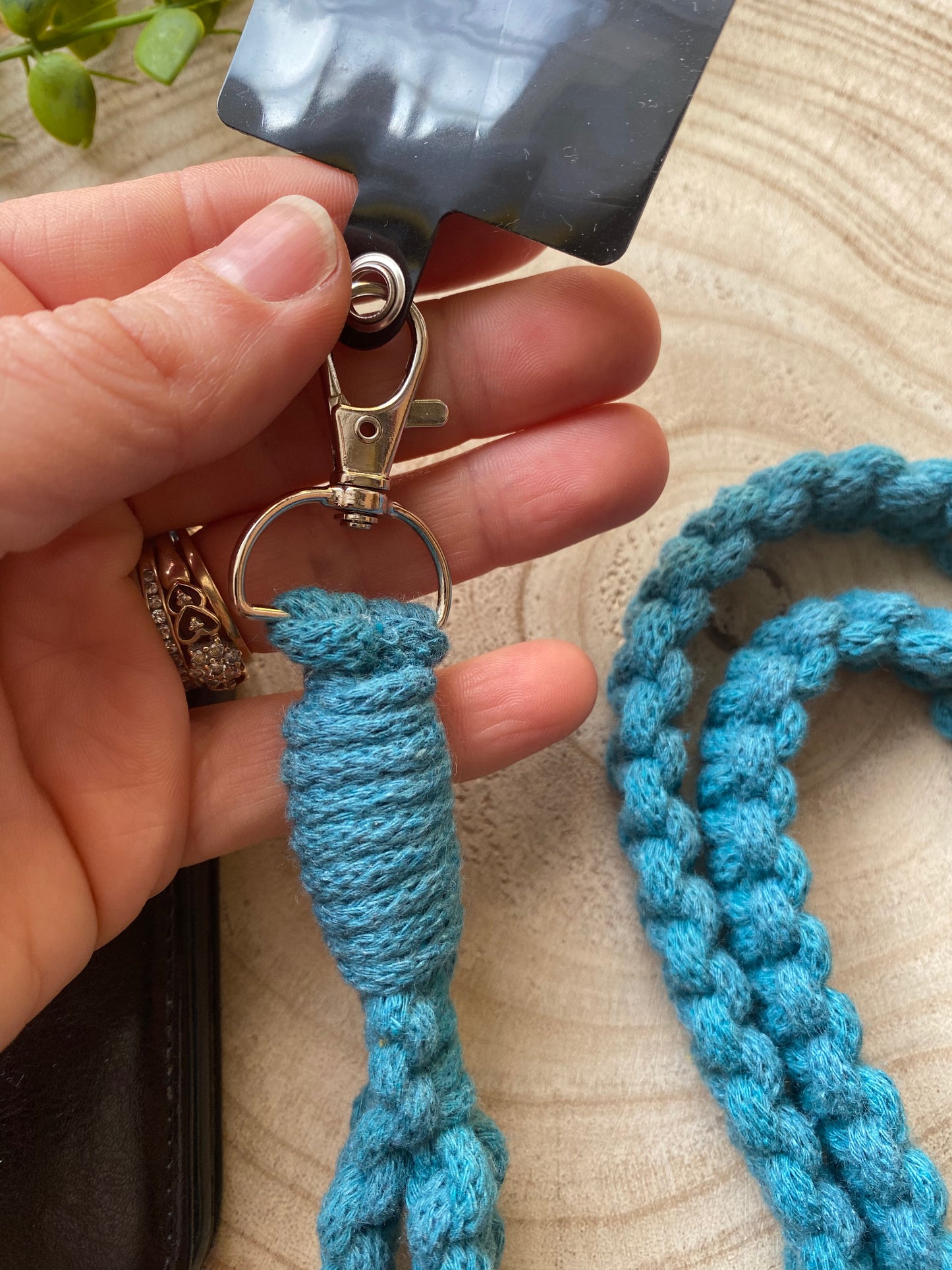 A hand holds a silver clasp attached to a thick, braided blue rope, revealing the Recycled Cotton Crossbody Phone Strap by Macra-Made-With-Love. There is a silver ring and a black badge holder at the end of the clasp. A small plant is visible in the background on a light wooden surface while adorned with rings, including a heart-shaped one.