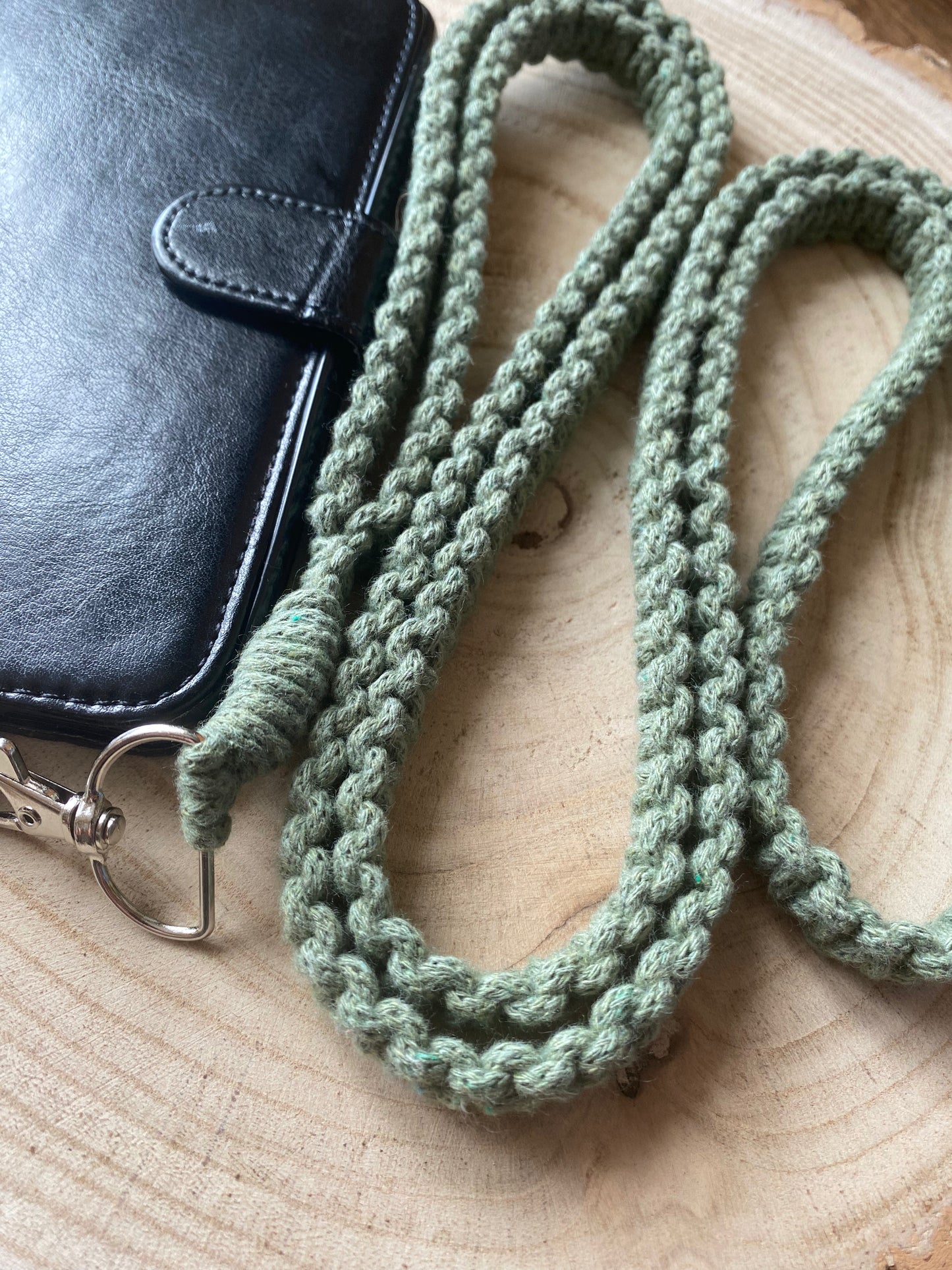 A black wallet with a strap sits on a wooden surface next to an eco-friendly, green, braided Recycled cotton crossbody phone strap by Macra-Made-With-Love.