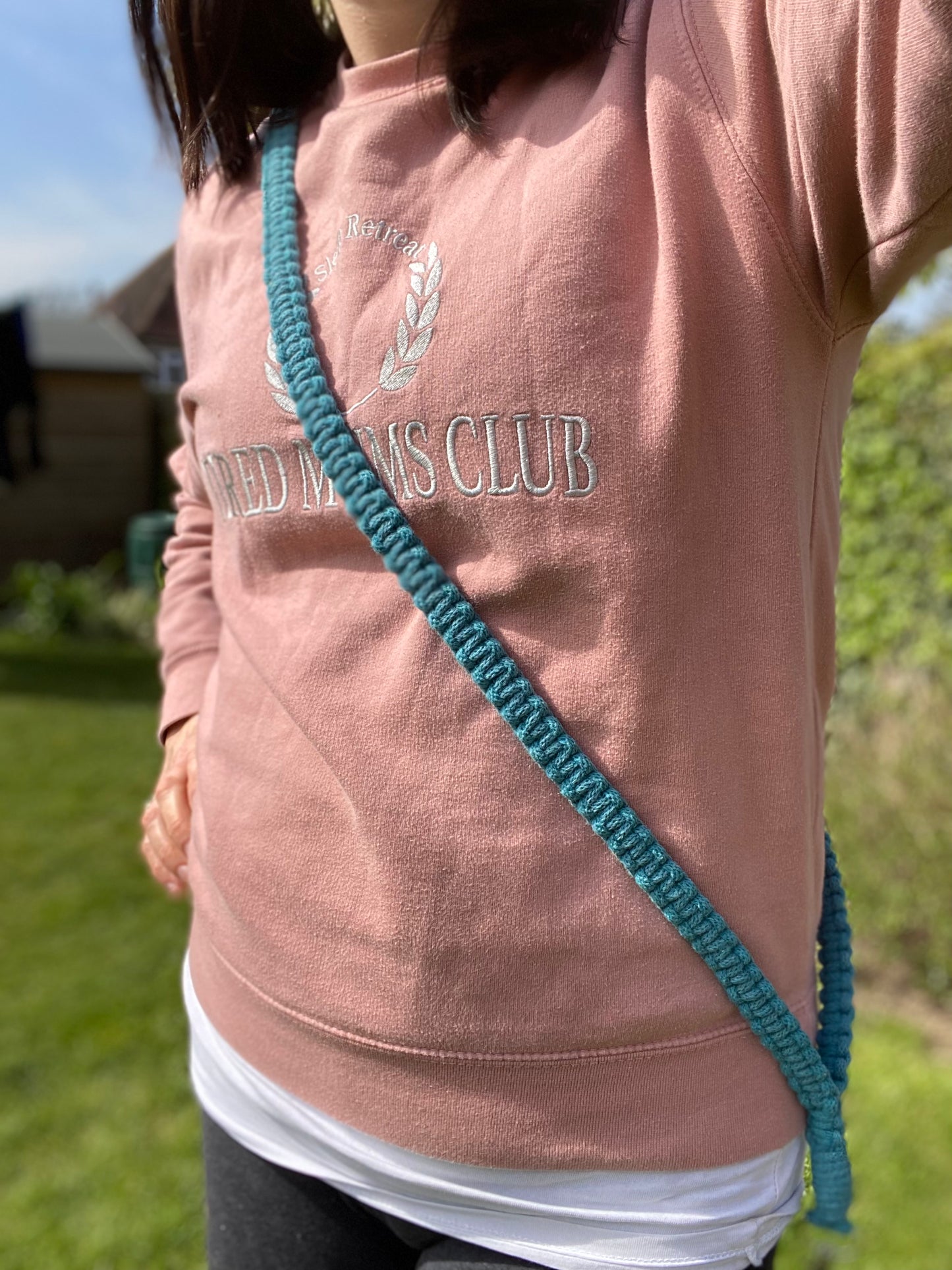 A person wearing a pink sweatshirt with the text "Bird Mens Club" and a green, handmade Macra-Made-With-Love Recycled cotton crossbody phone strap across their chest stands outdoors on a sunny day. The background includes green grass and some trees or bushes.