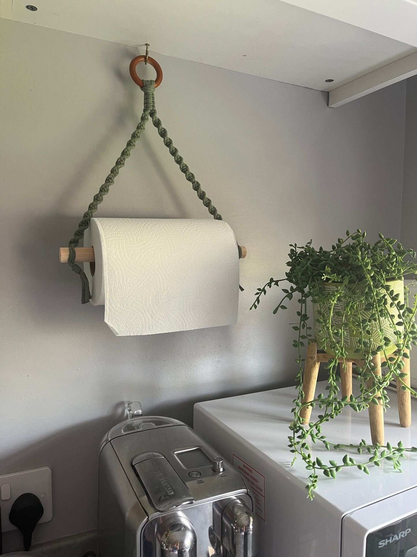 Roll of white paper towel on a macrame wall hanging holder made with sage green yarn and a wooden ring, next to a plant which is placed on top of a white microwave.