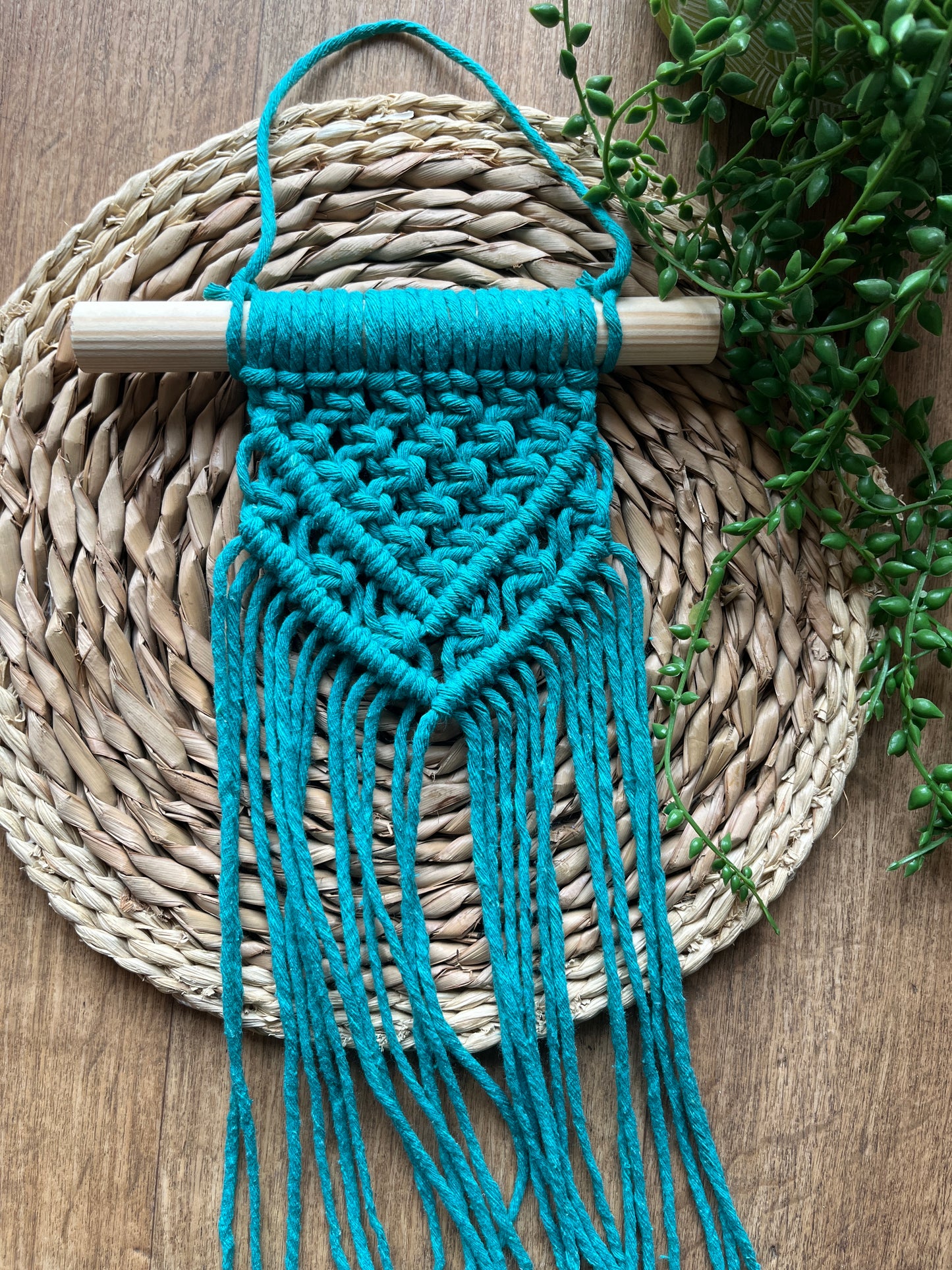 Blue macrame wall hanging on a wooden dowel, laid across a round woven mat with greenery