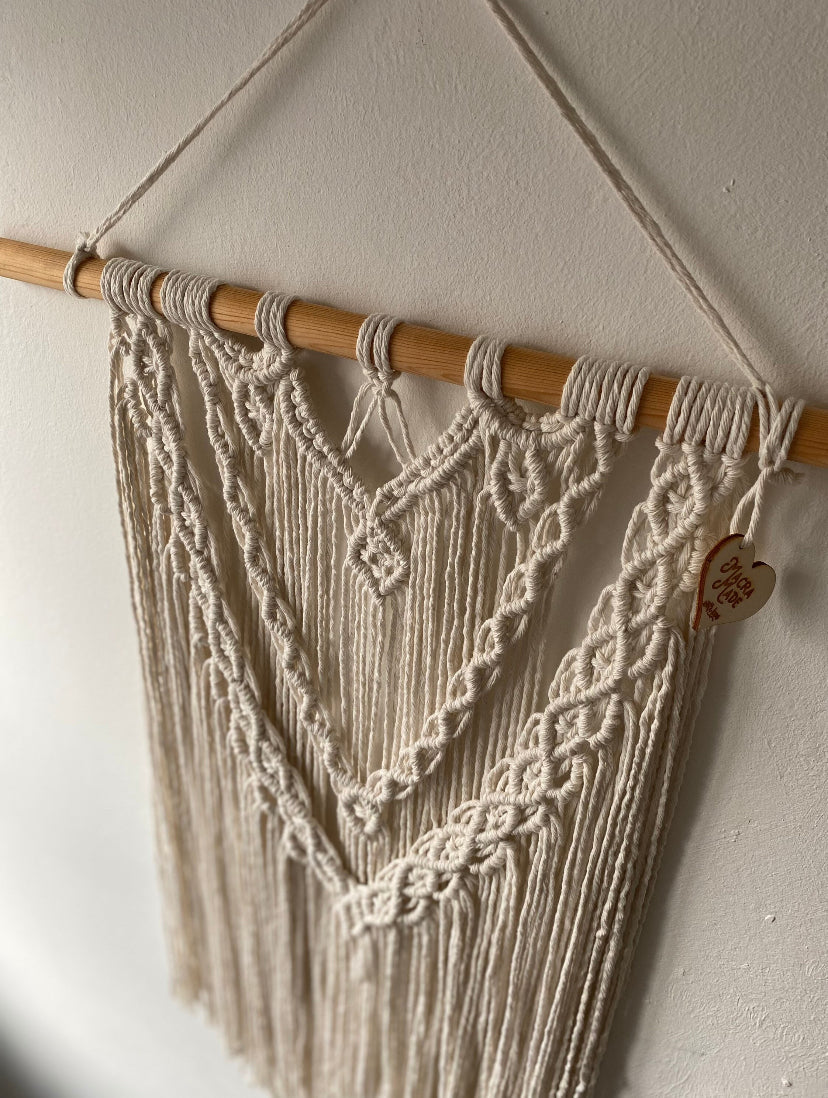 A top down view of a large statement macrame wall hanging made with natural cotton yarn on a long wooden dowel, with layered knots and boho style. Hanging on a white wall.