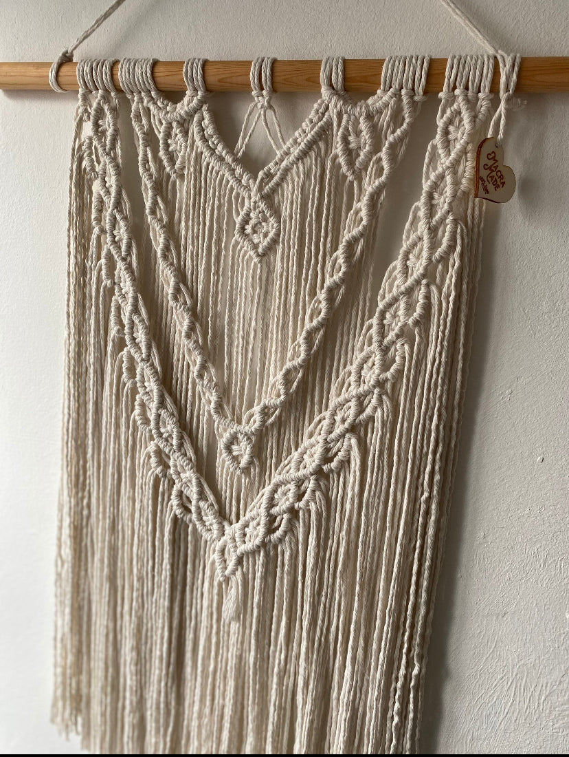 A close up of a large statement macrame wall hanging made with natural cotton yarn on a long wooden dowel, with layered knots and boho style. Hanging on a white wall.