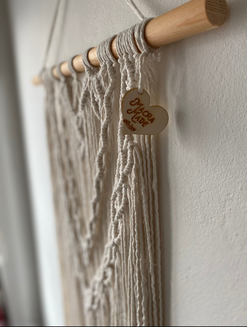 Close up side angle of a large statement macrame wall hanging made with natural cotton yarn on a long wooden dowel, with layered knots and boho style. Hanging on a white wall.