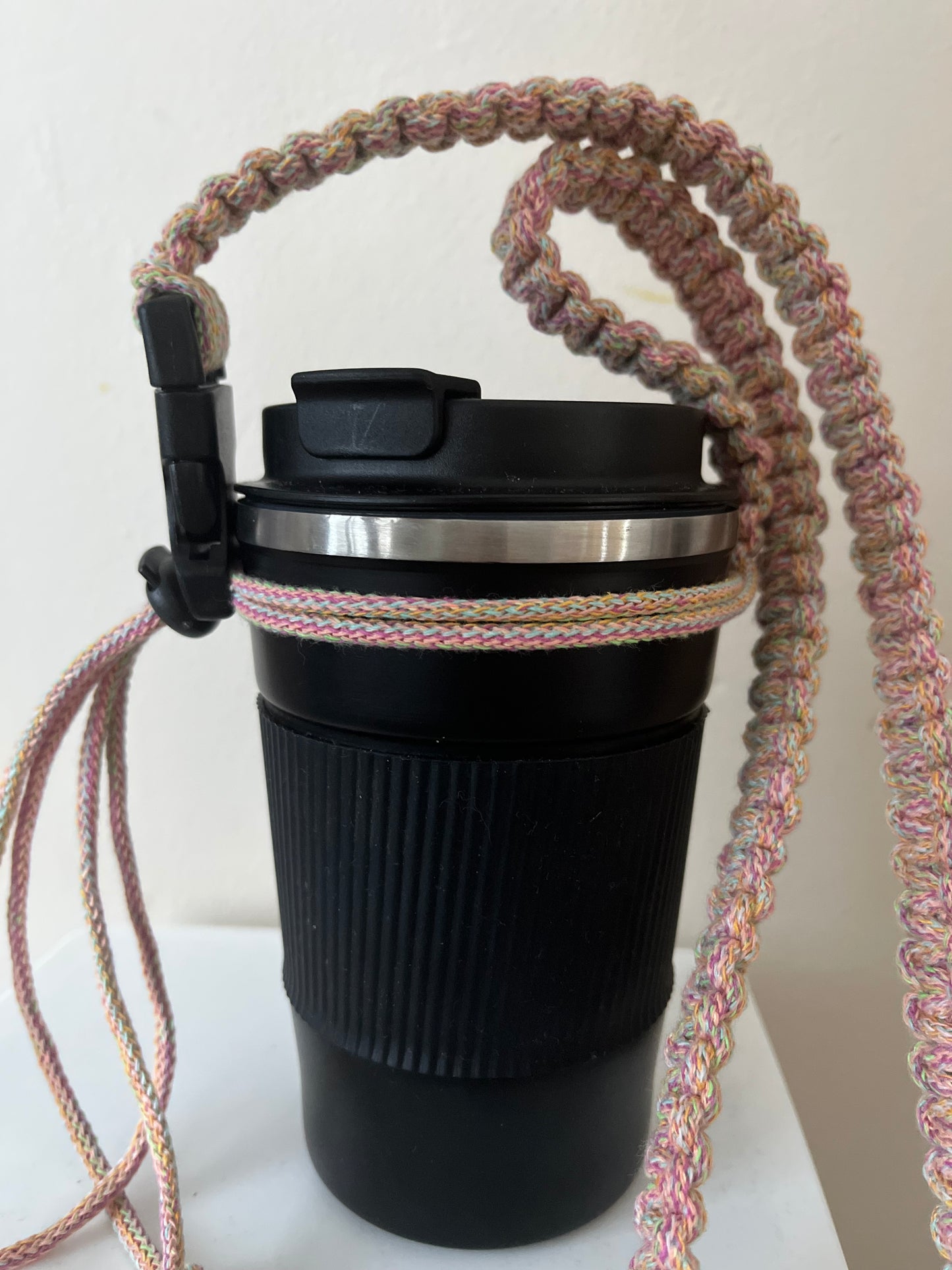 A black travel cup stands in front of a  white wall, with a long rainbow coloured macrame strap attached to it. The strap features neat knots and has a black buckle and toggle.