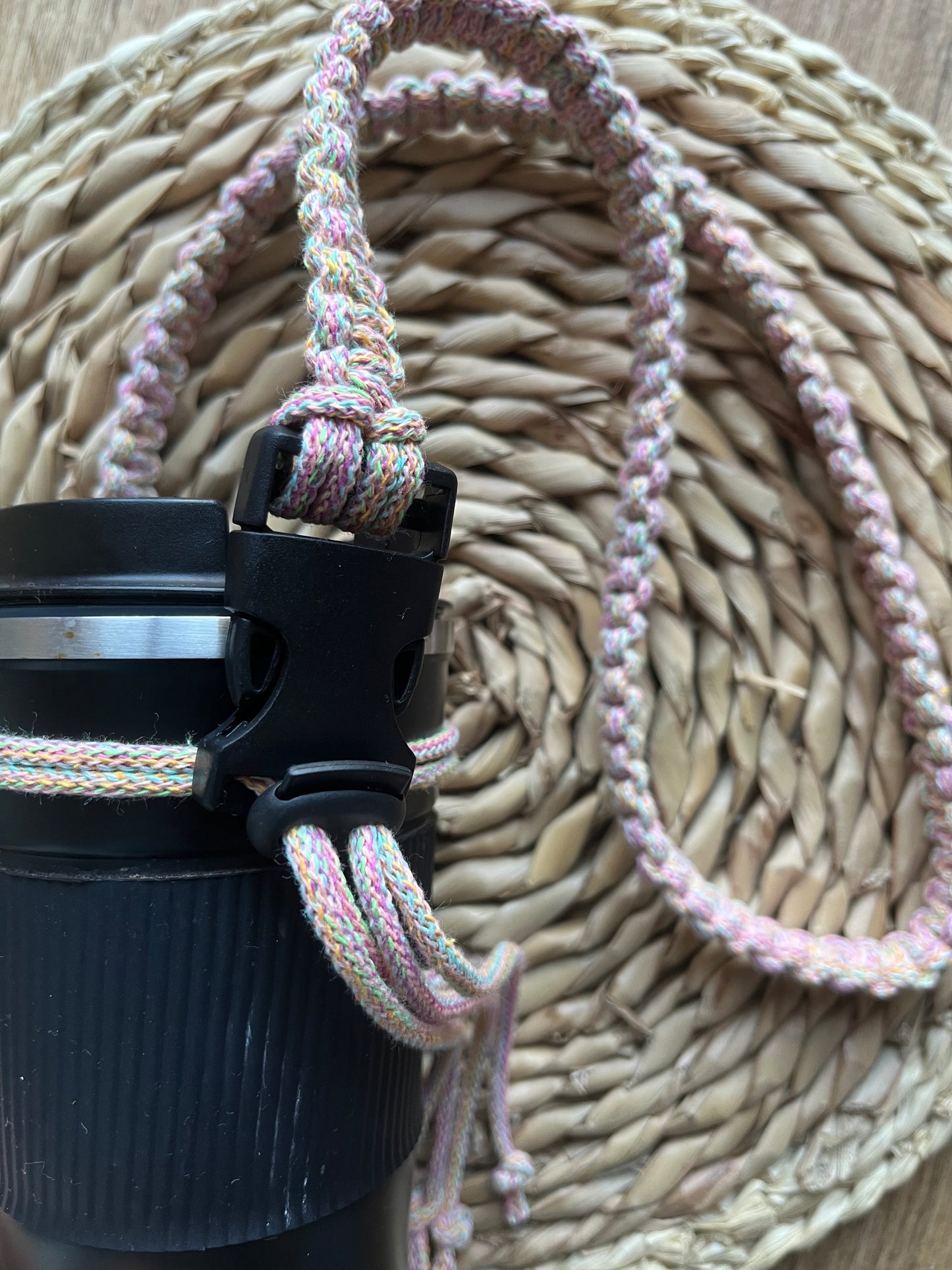 A black reusable travel cup is laying on a wicker mat with a handmade macrame strap attached to it, the strap is made with rainbow coloured recycled cotton, with a black toggle and buckle.