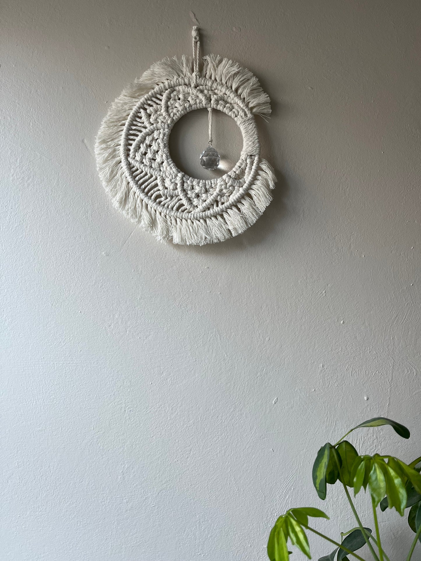 Handmade macrame moon shaped boho sun catcher made with recycled cotton natural yarn and a crystal prism in the centre. Displayed hanging on a plain painted wall, with a green plant just visible in the bottom right corner.