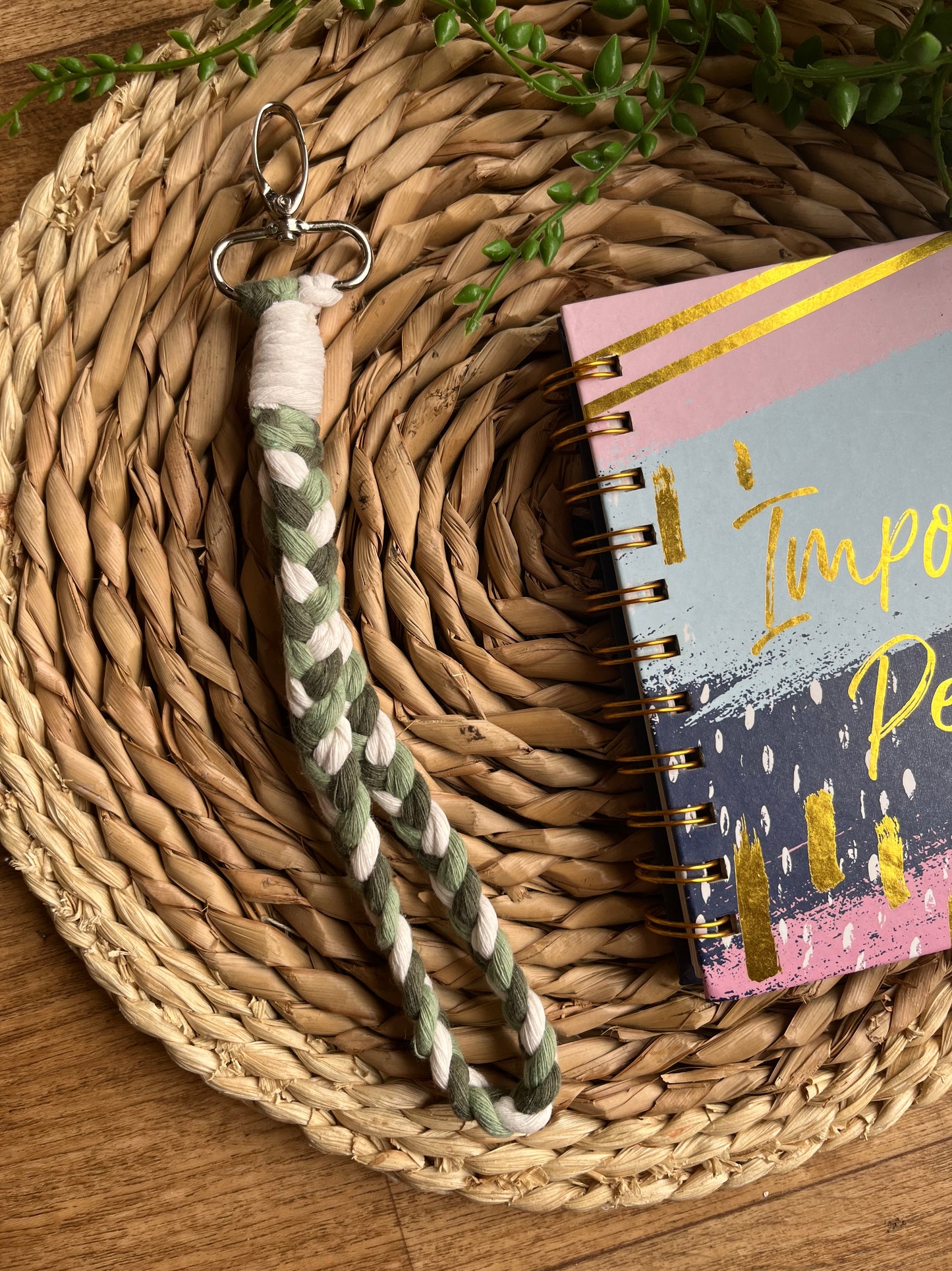 A photo of a handmade macrame braided wristlet, laid on a wicker mat next to a notebook. The wristlet is made with green and natural recycled cotton, with a metal lobster clasp.