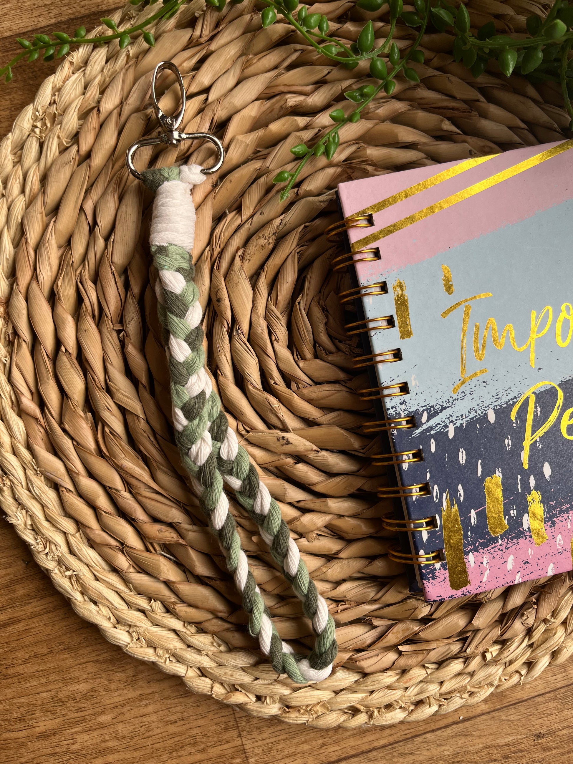 A photo of a handmade macrame braided wristlet, laid on a wicker mat next to a notebook. The wristlet is made with green and natural recycled cotton, with a metal lobster clasp.