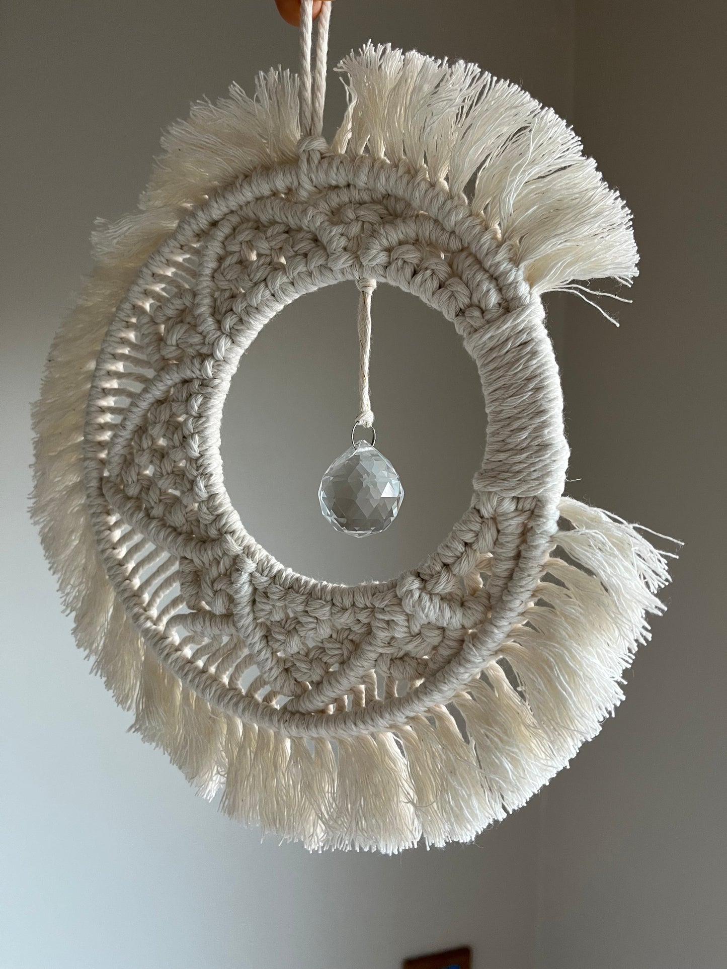 Handmade macrame moon shaped boho sun catcher made with recycled cotton natural yarn and a crystal prism in the centre. Displayed hanging in front of a plain painted background.