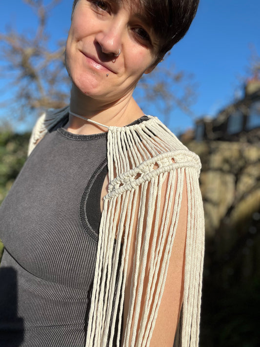 A female wearing a black vest, and handmade macrame shoulder accessory epaulette, stands outside on a sunny day, smiling towards the camera.