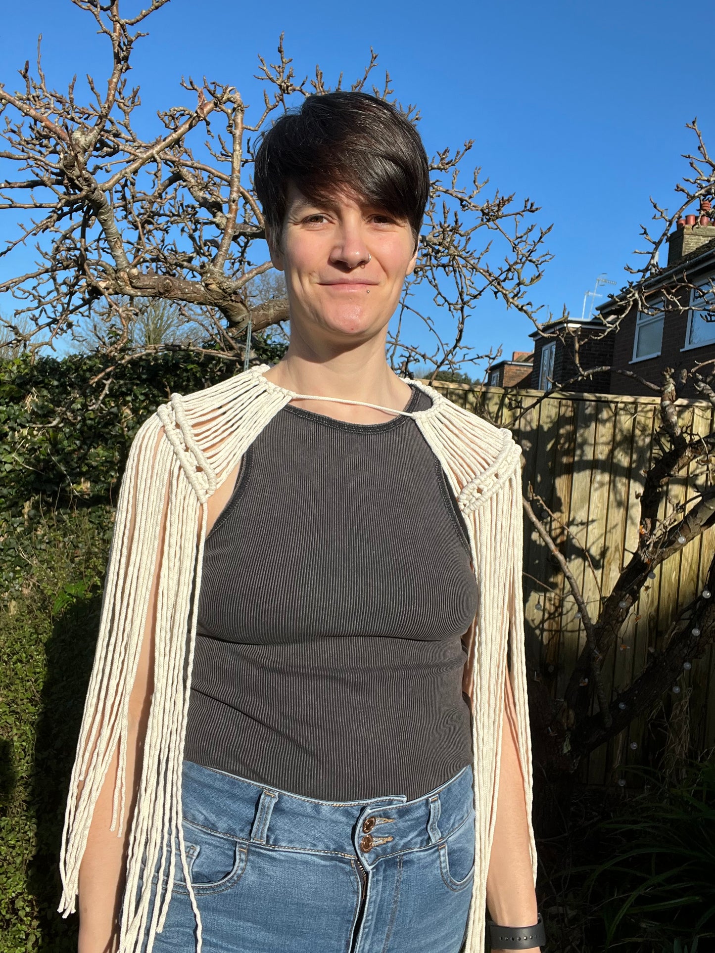 A female stands outside on a sunny day wearing black vest, blue jeans and a handmade macrame shoulder accessory epaulette made with natural recycled cotton. She is looking at the camera and smiling.